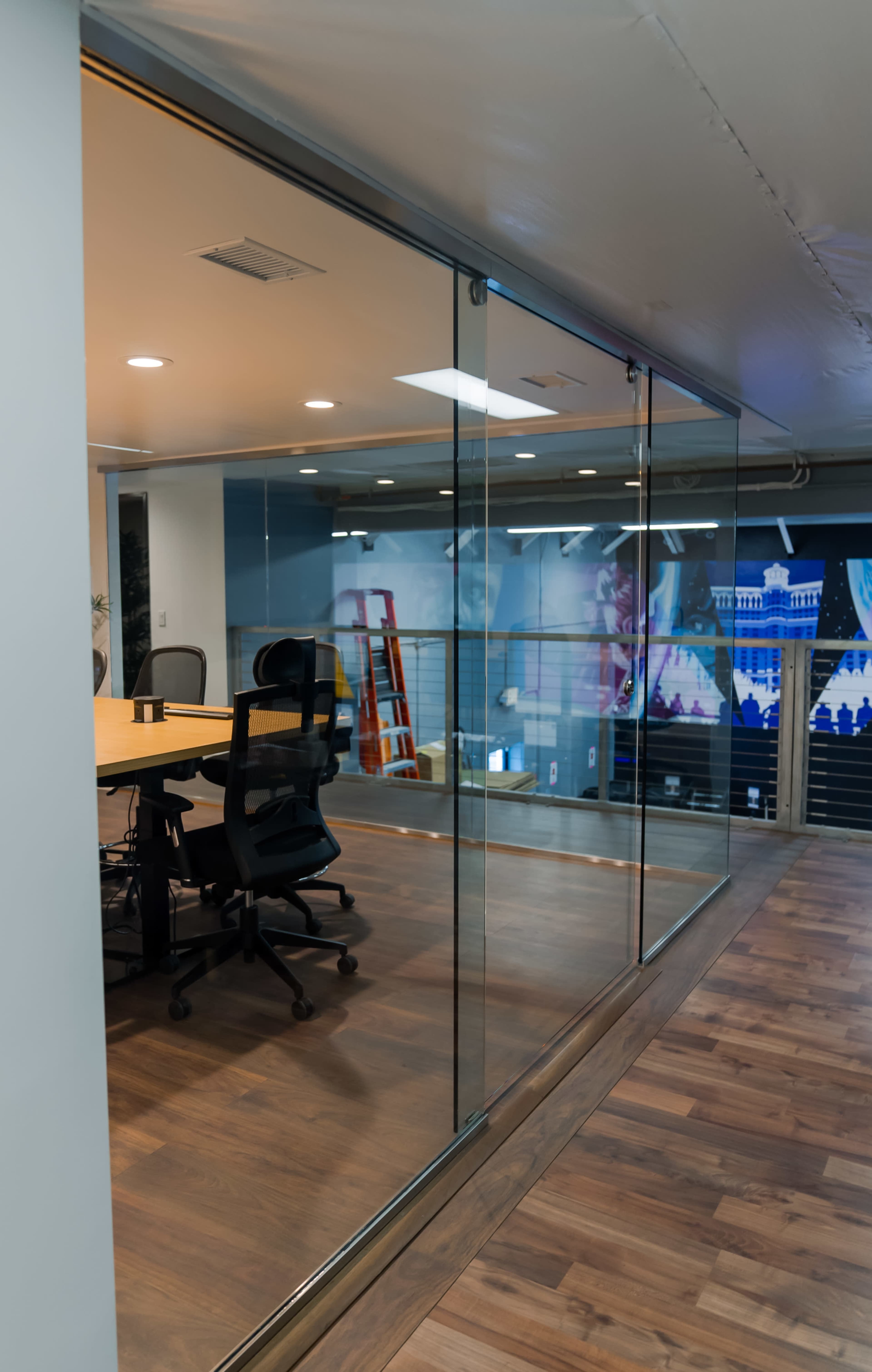 A modern office space features a glass-walled conference room with a round table and rolling chairs, visible alongside a mural and a ladder in the background.
