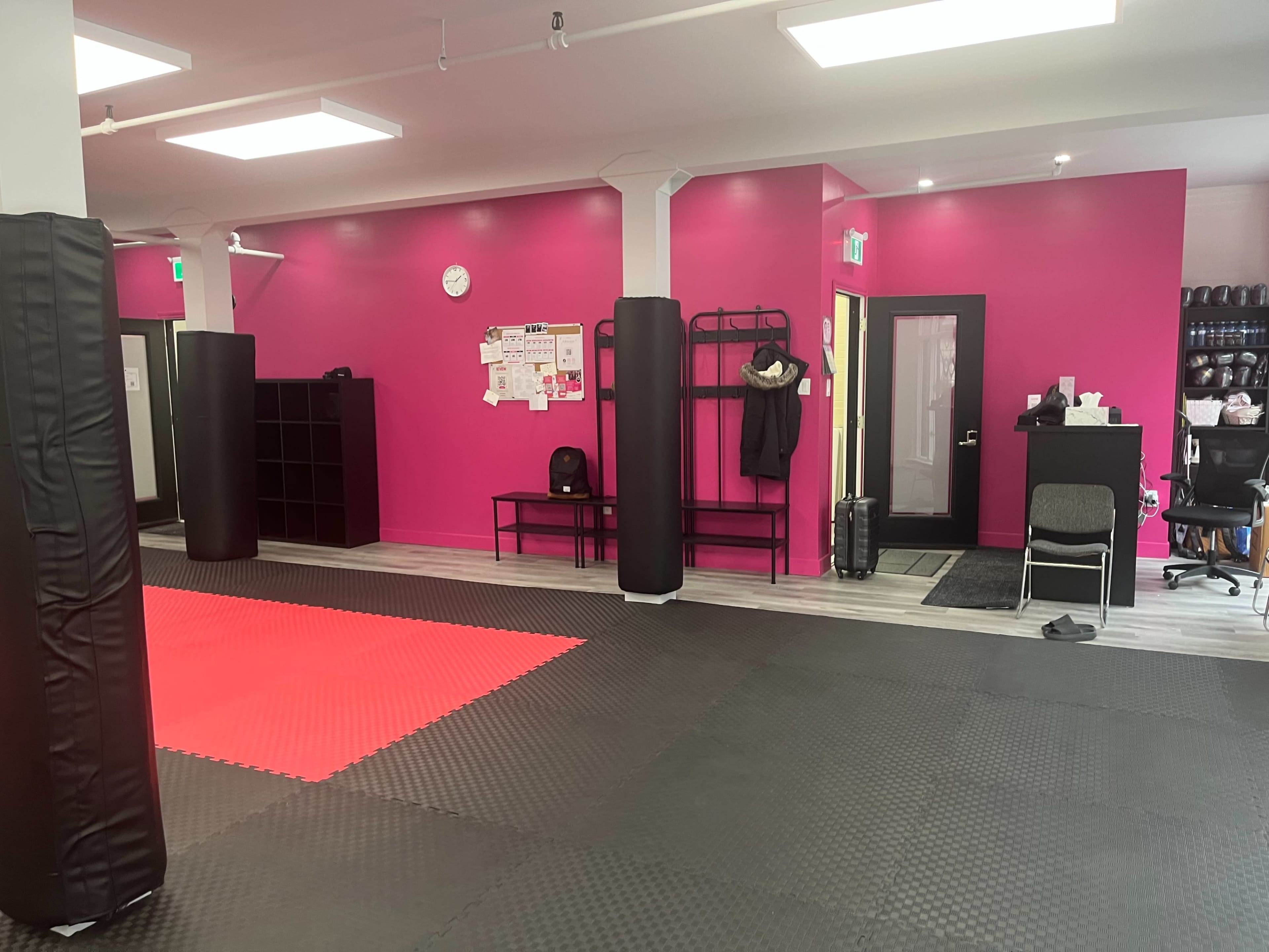 The image shows a martial arts studio with a red padded training area, black wall pads, and vibrant pink walls, along with equipment and seating areas in the background.