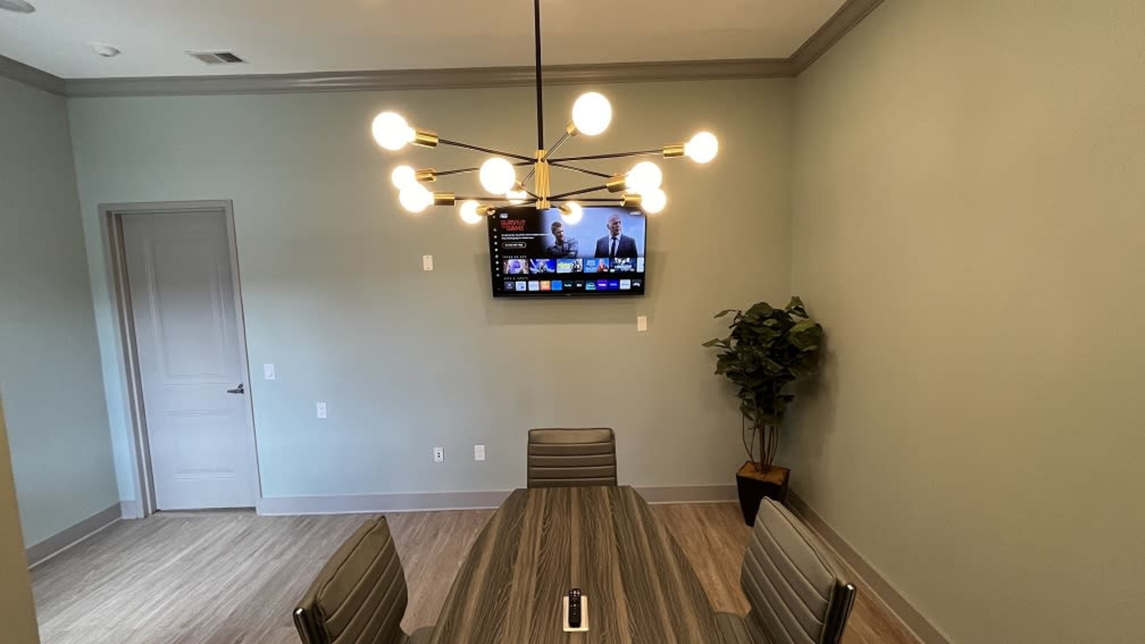 A modern conference room with a table surrounded by chairs, a wall-mounted television, and a potted plant in the corner.