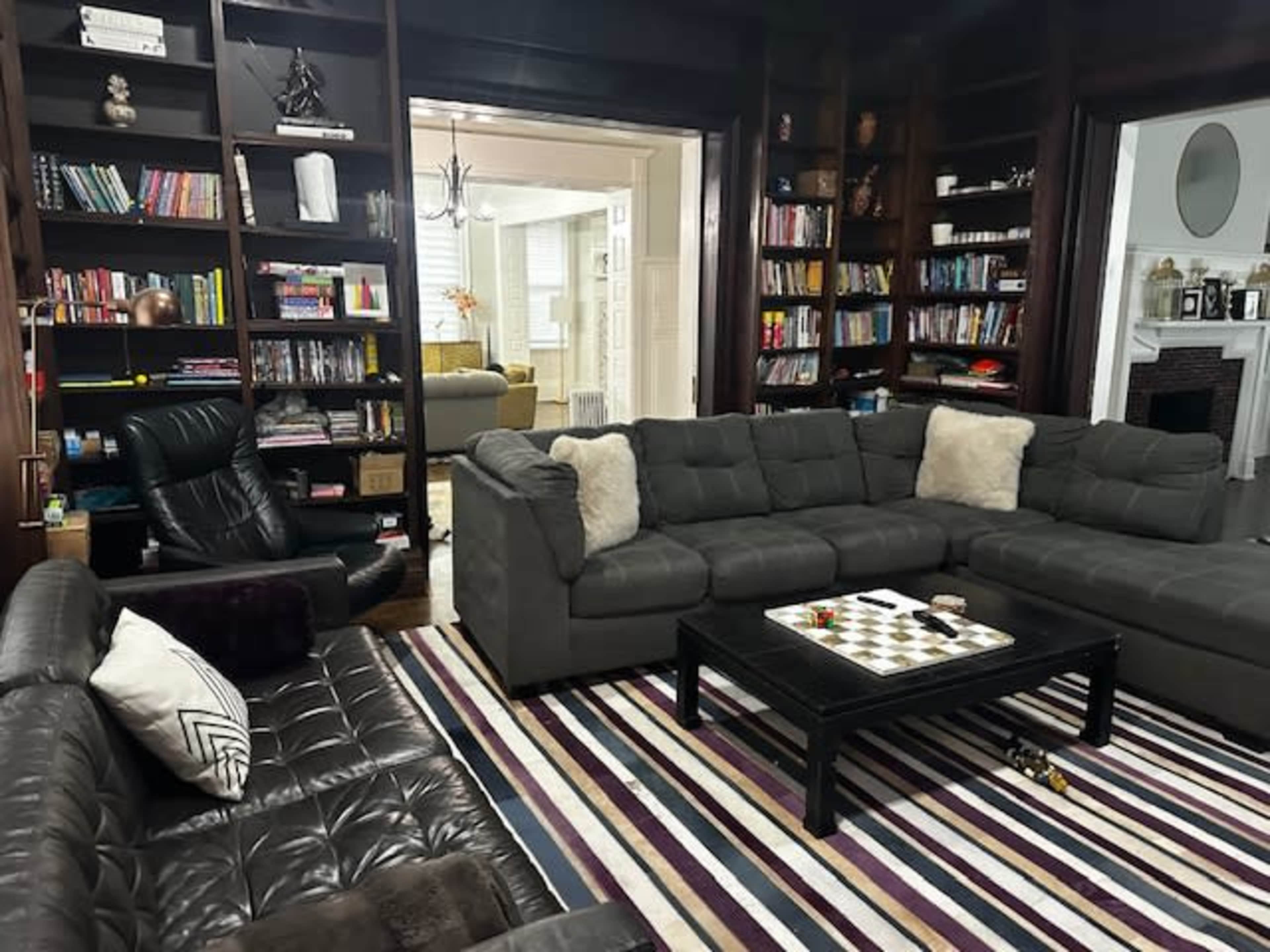 The image shows a living room with two large sectional sofas, a coffee table with a chessboard, and a bookcase filled with books in the background.