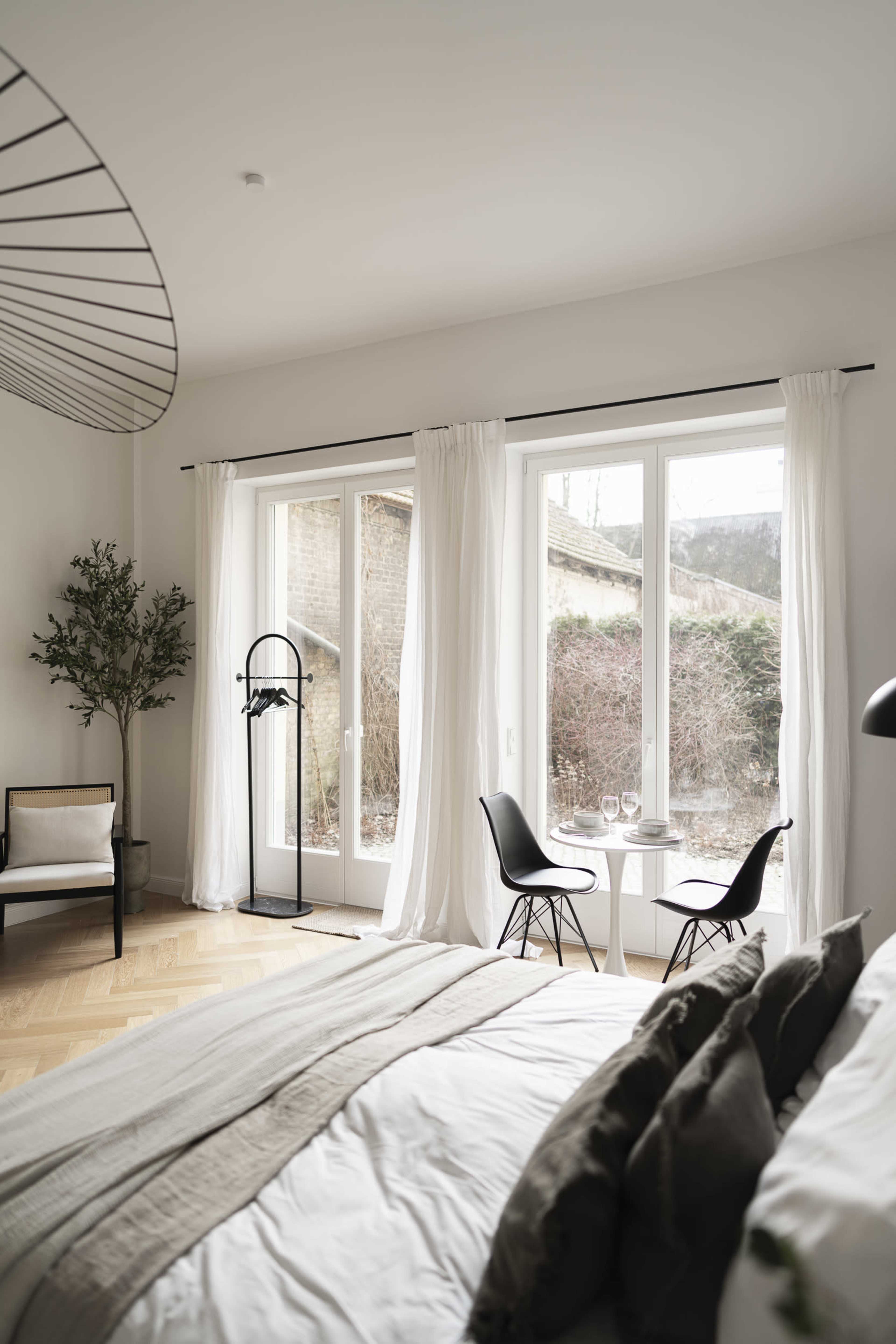 A spacious bedroom features a bed with neutral linens, two black chairs around a small table, and large windows that overlook a garden.