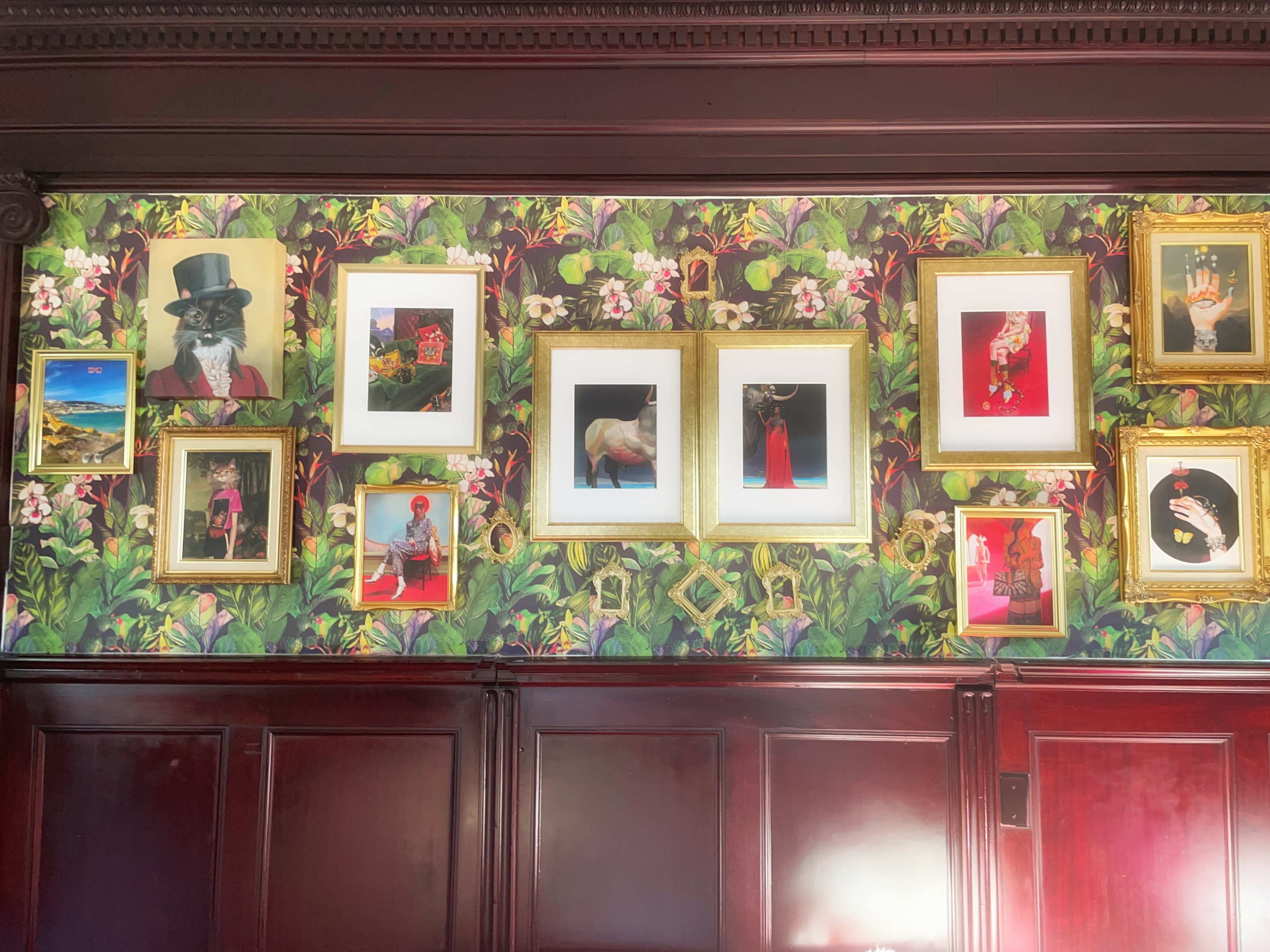 A wall covered in vibrant floral wallpaper adorned with various framed artworks, including portraits and photographs in gold and white frames.