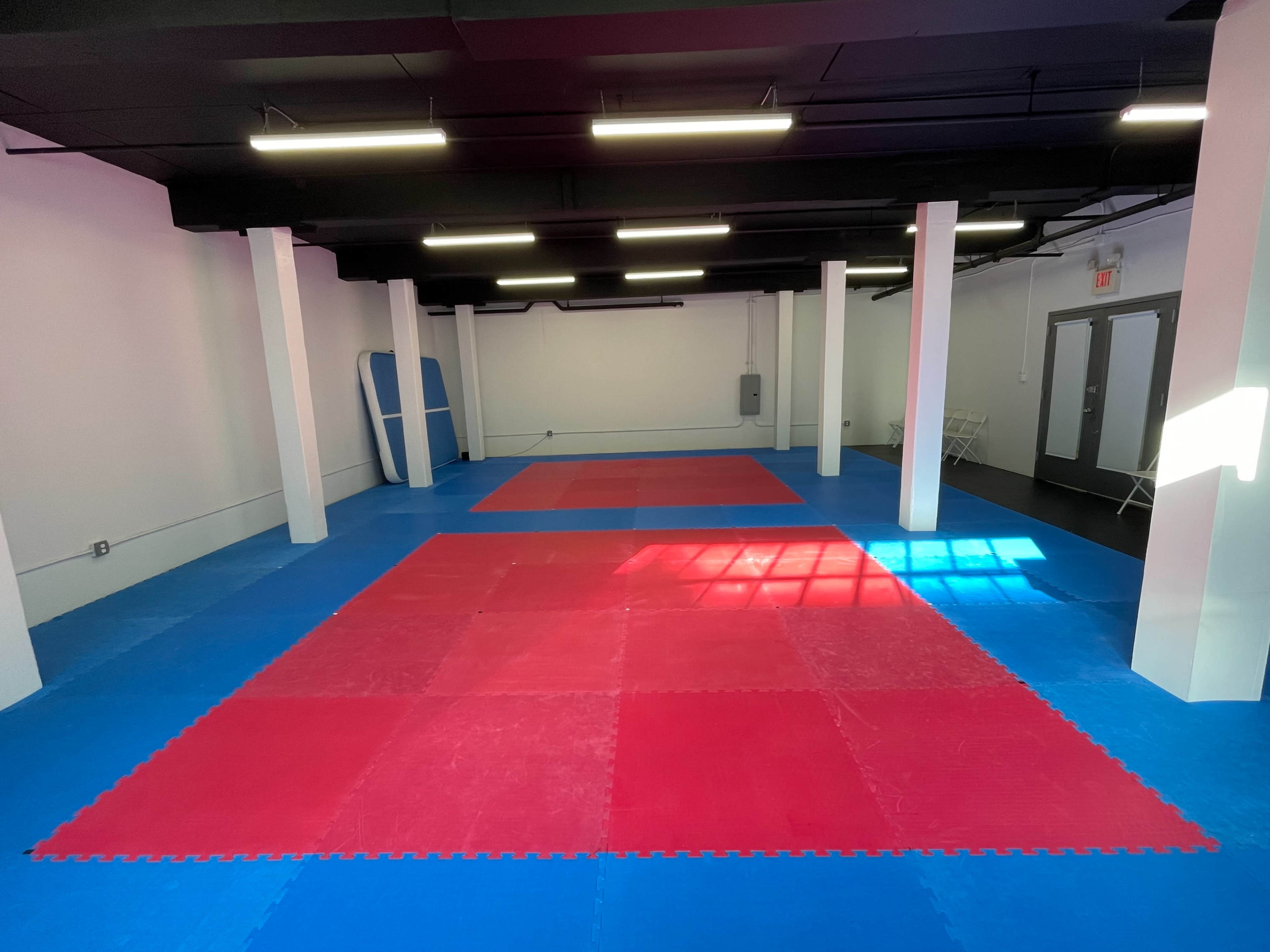 A spacious martial arts studio with red and blue puzzle mats arranged on the floor, illuminated by fluorescent lights.