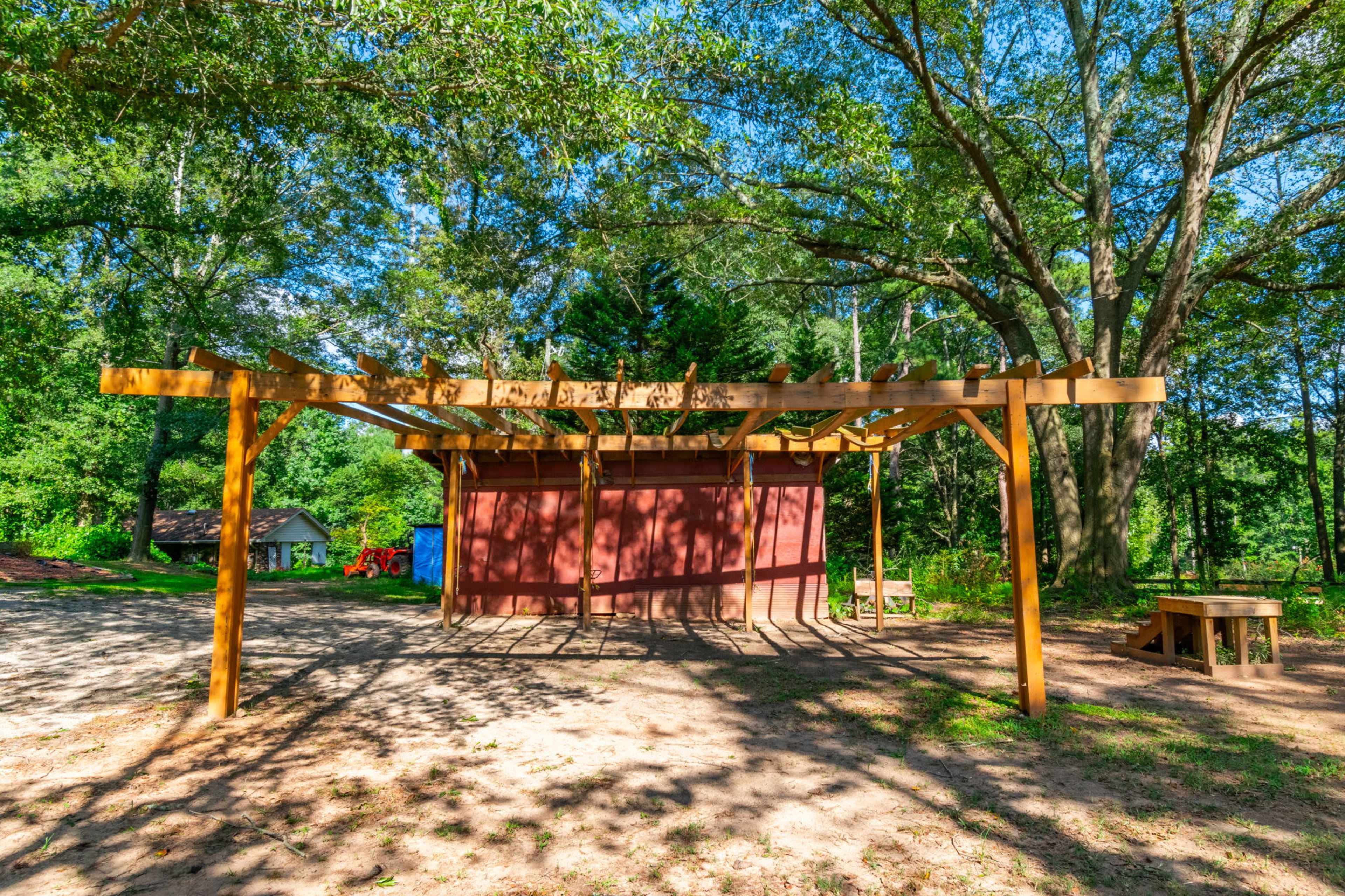 A wooden pergola stands beside a red storage shed in a clear, sunny outdoor area surrounded by trees.