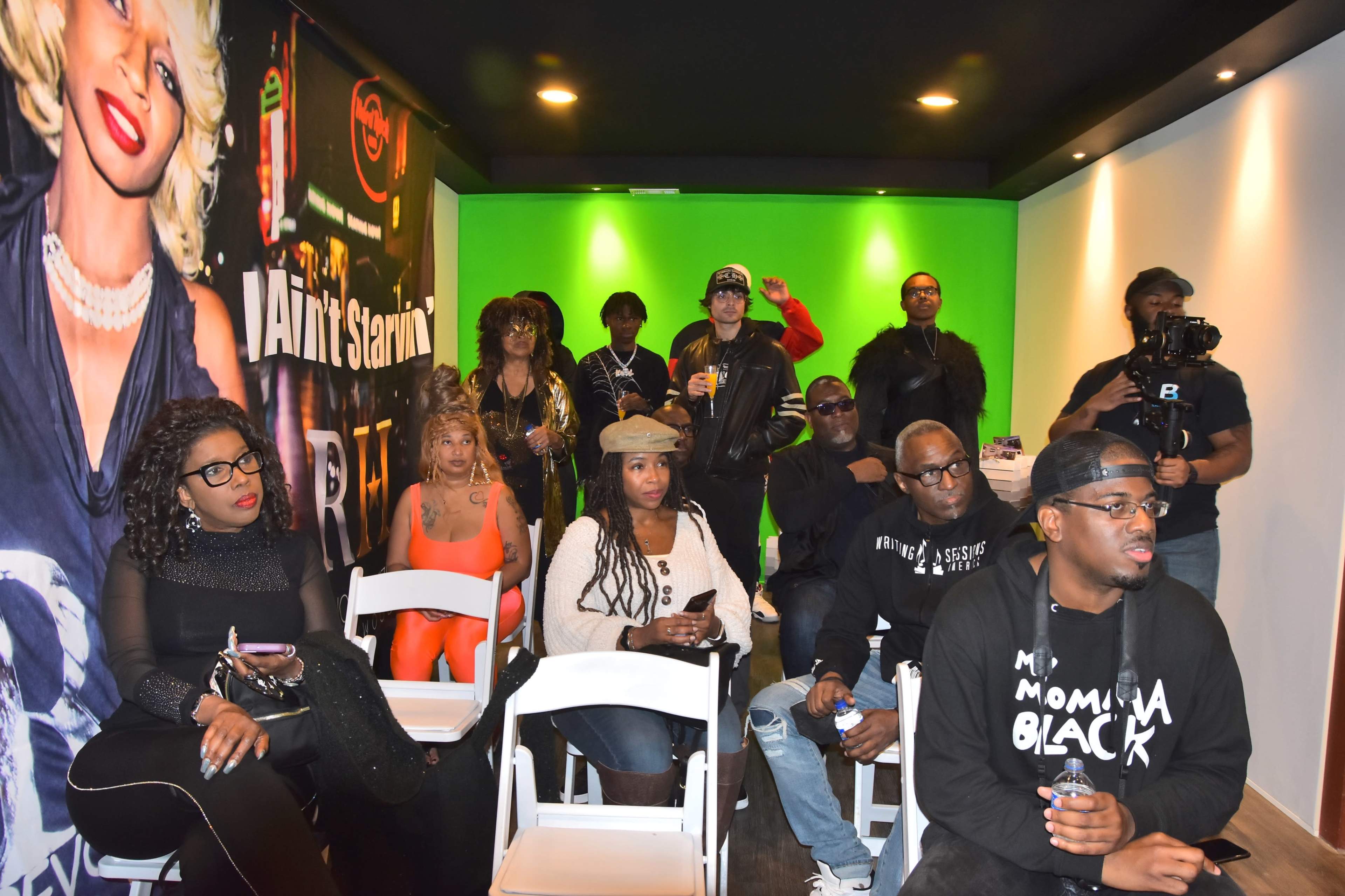 A diverse group of people sits in a room with a green screen backdrop, some facing the camera while others are engaged in conversation.