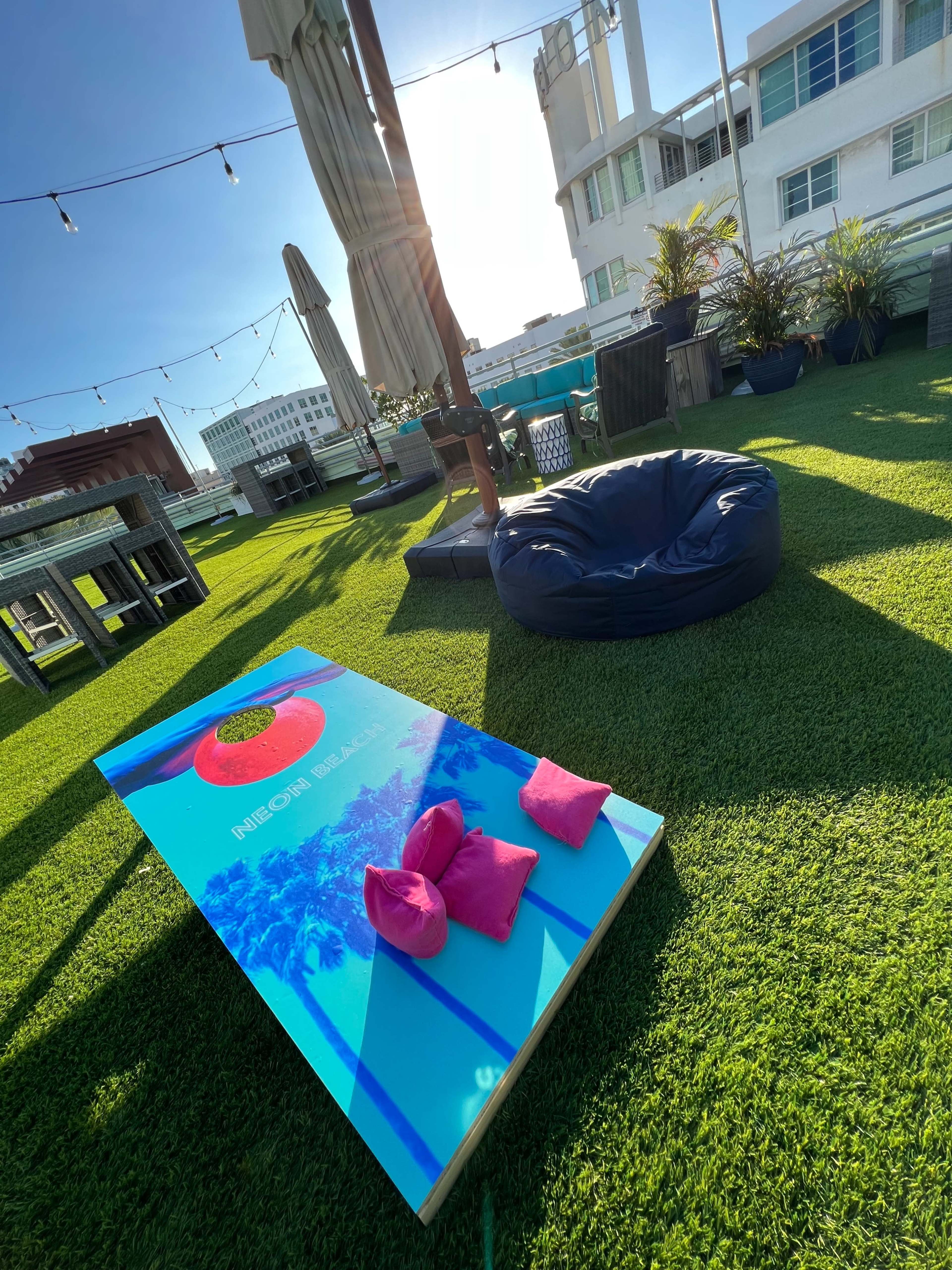 The image shows a rooftop space with artificial grass, featuring a bean bag chair, a cornhole game set, and several tall umbrellas.