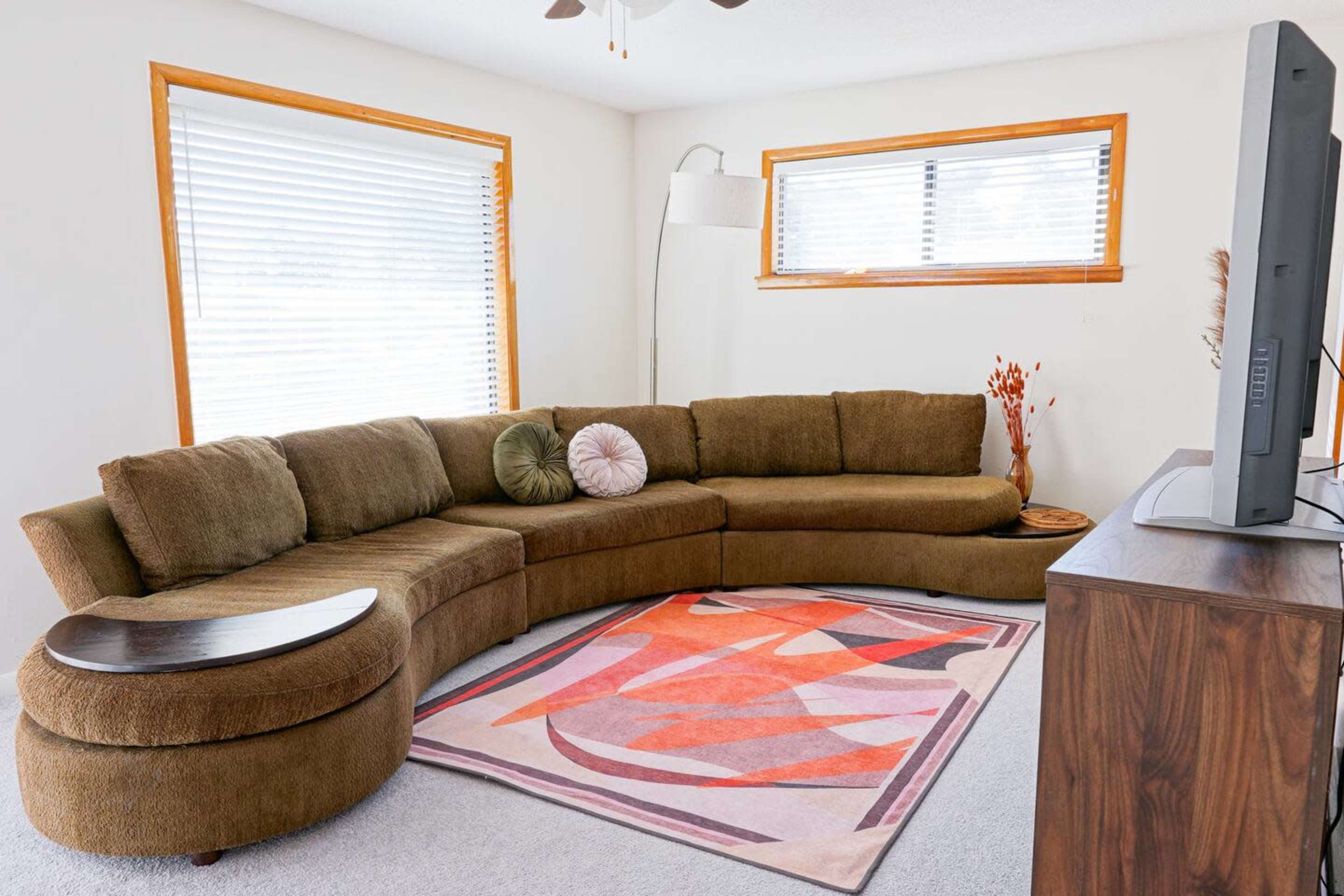 The image shows a living room with a large, curved brown sectional sofa, a light-colored area rug with geometric patterns, and a flat-screen television on a wooden stand.