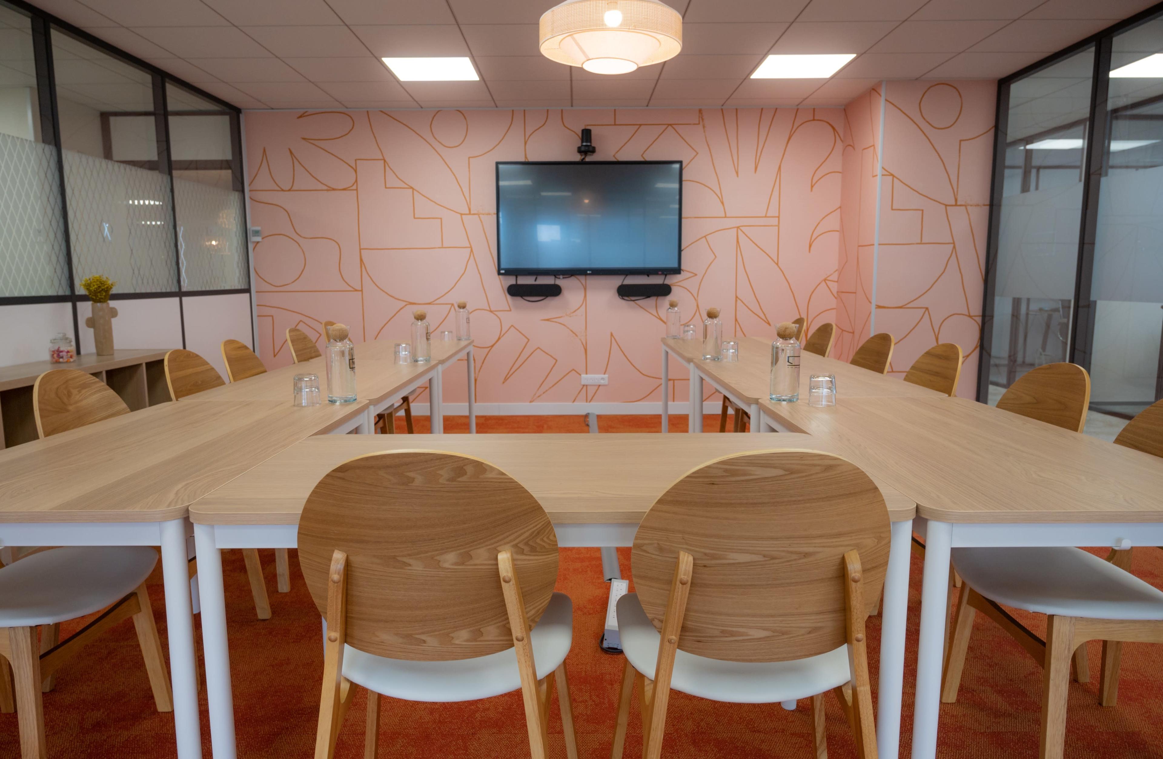 A meeting room features multiple wooden tables arranged in a U-shape, surrounded by chairs, with a large screen displaying on the wall.