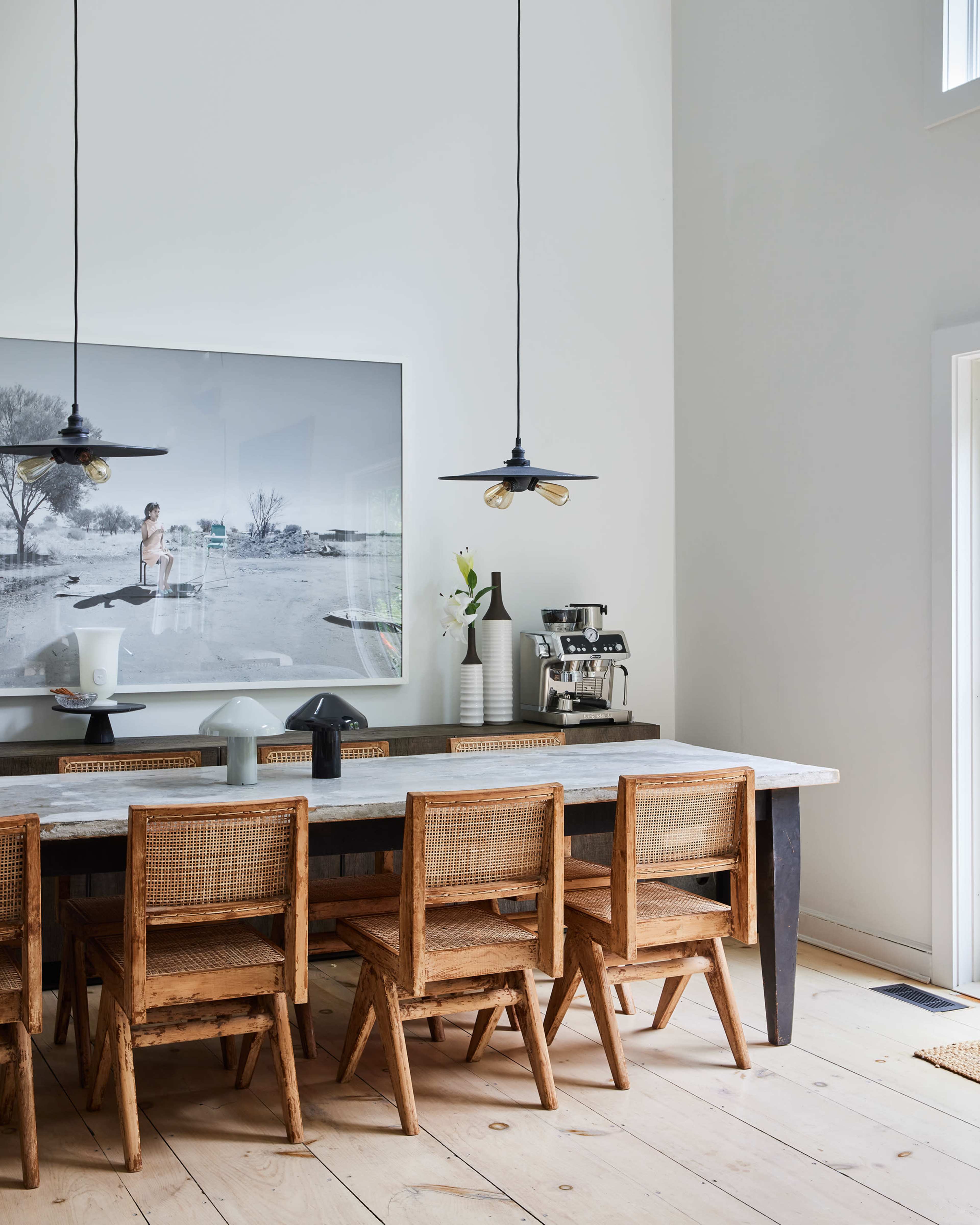 A sleek dining area features a long wooden table with several woven chairs and two pendant lights hanging above, complemented by a large black-and-white photograph on the wall.