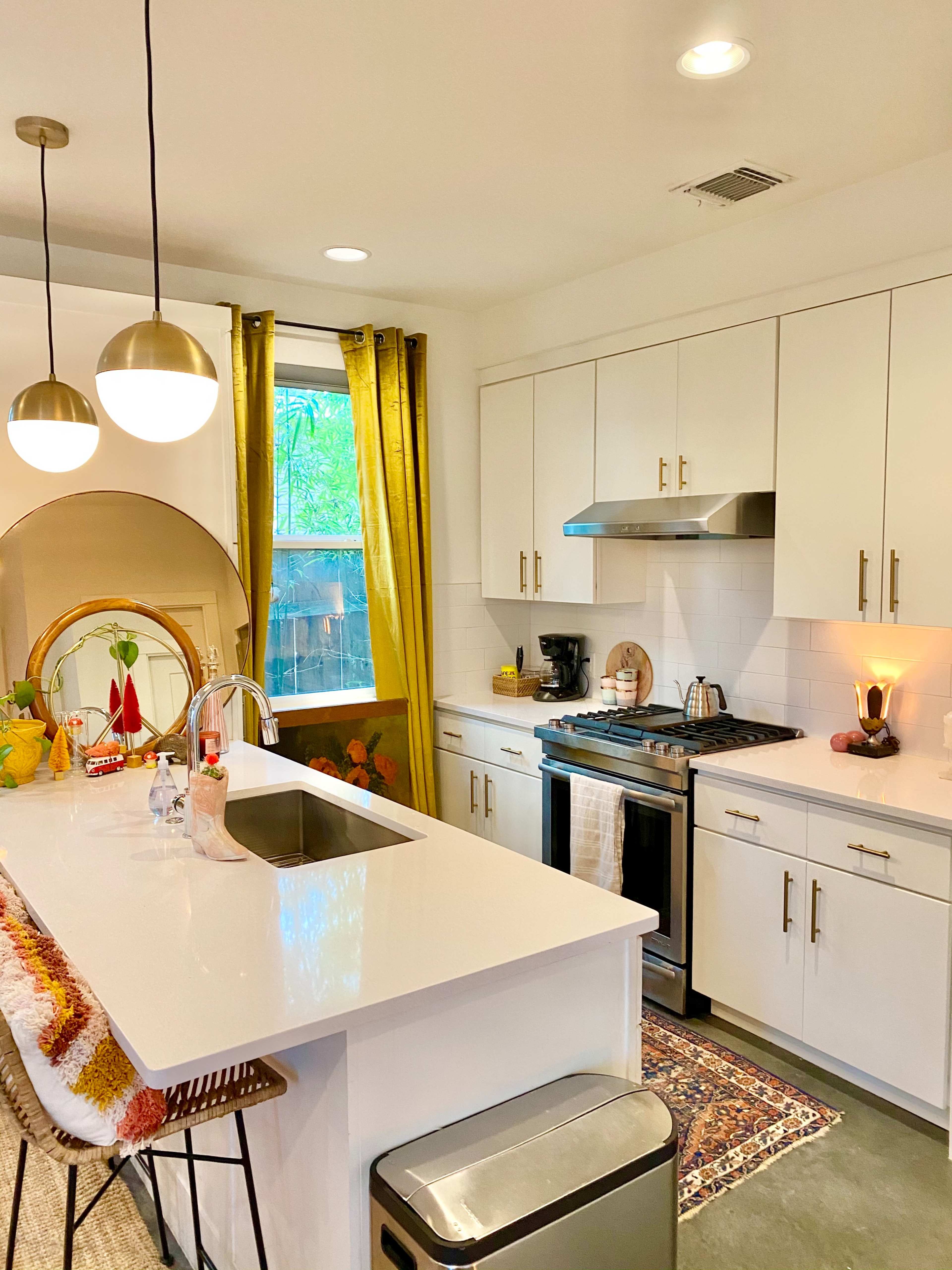 A modern kitchen features white cabinets, a stainless steel stove, a large island with bar stools, and yellow curtains framing a window.