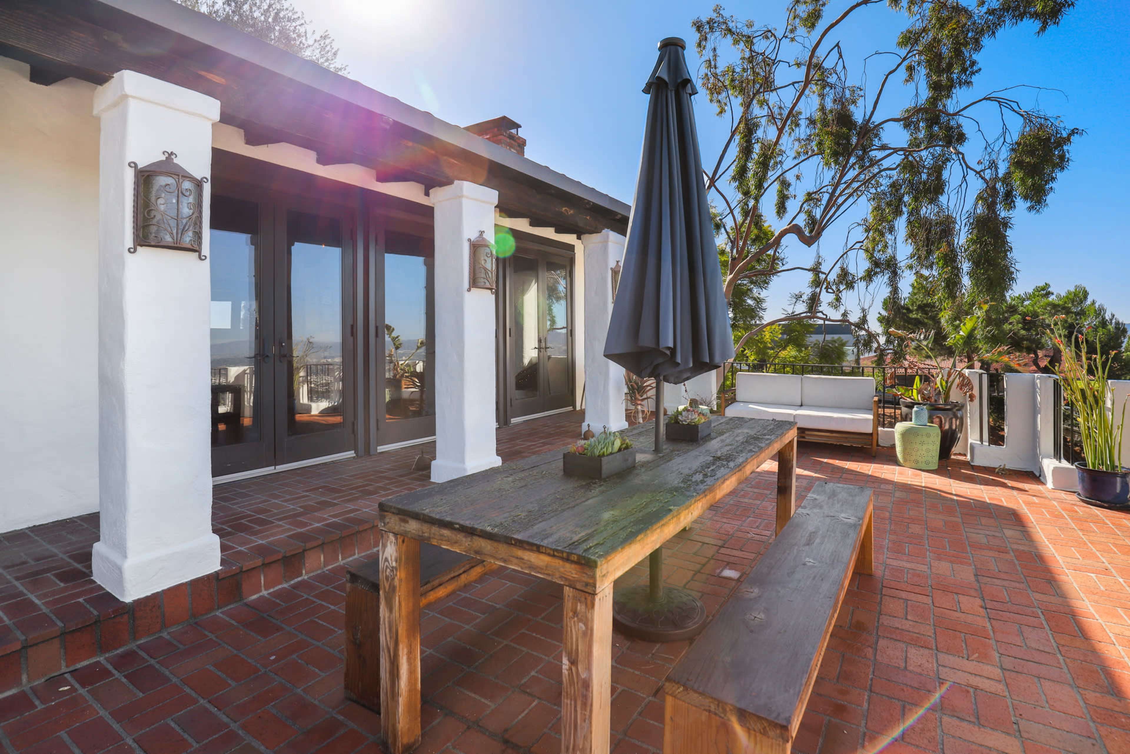 A wooden dining table with a bench is positioned on a brick patio, accompanied by a large umbrella and plants, with a view of the sky and greenery.