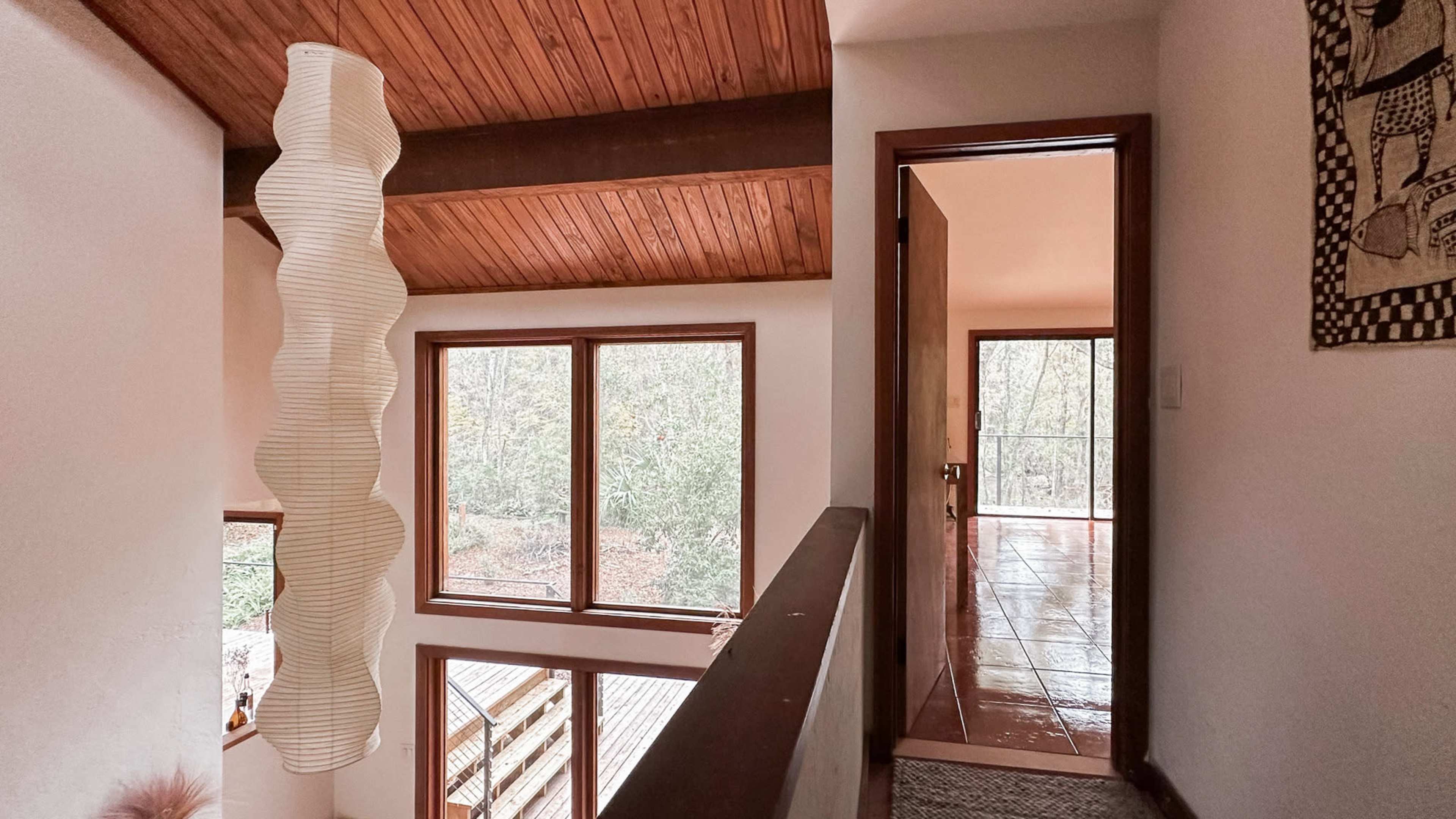 The image shows a hallway with a wooden ceiling, large windows overlooking greenery, and a hanging light fixture.