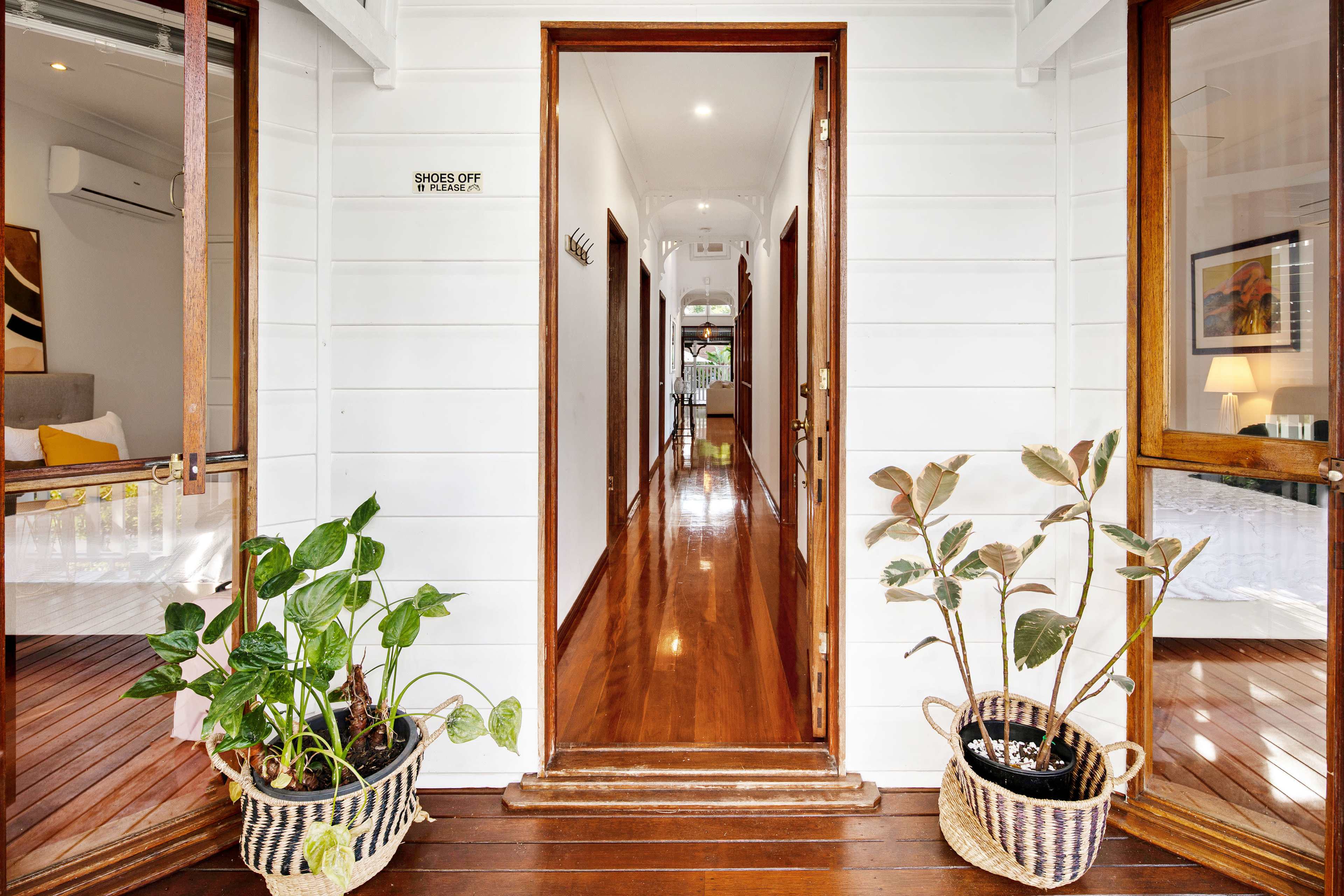 The entrance of a house with a wooden floor, two potted plants on either side of the doorway, and a hallway visible in the background leading to a light-filled room.
