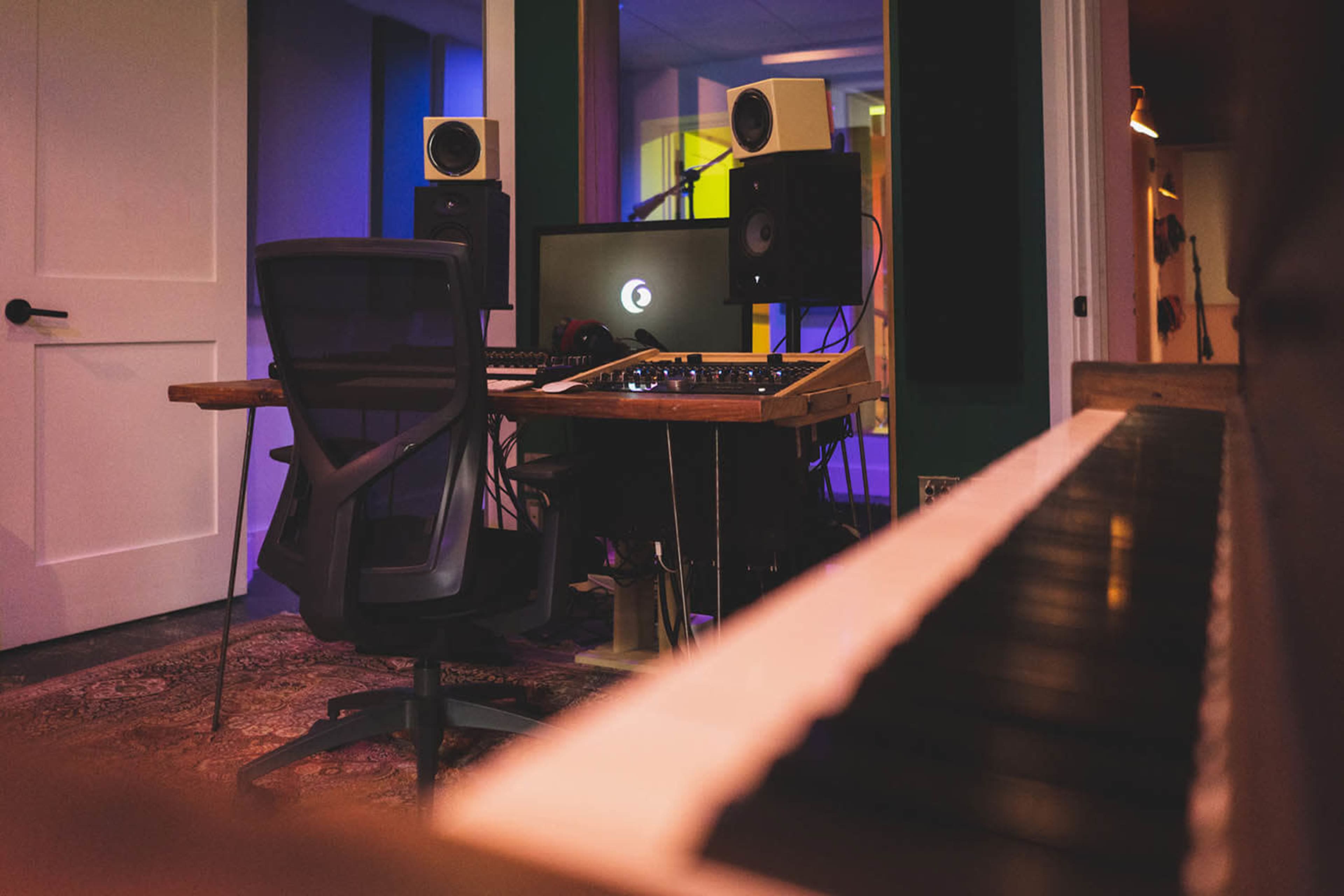 A recording studio with a mixing console, speakers, and a computer setup, viewed from an angle that includes a piano keyboard in the foreground.
