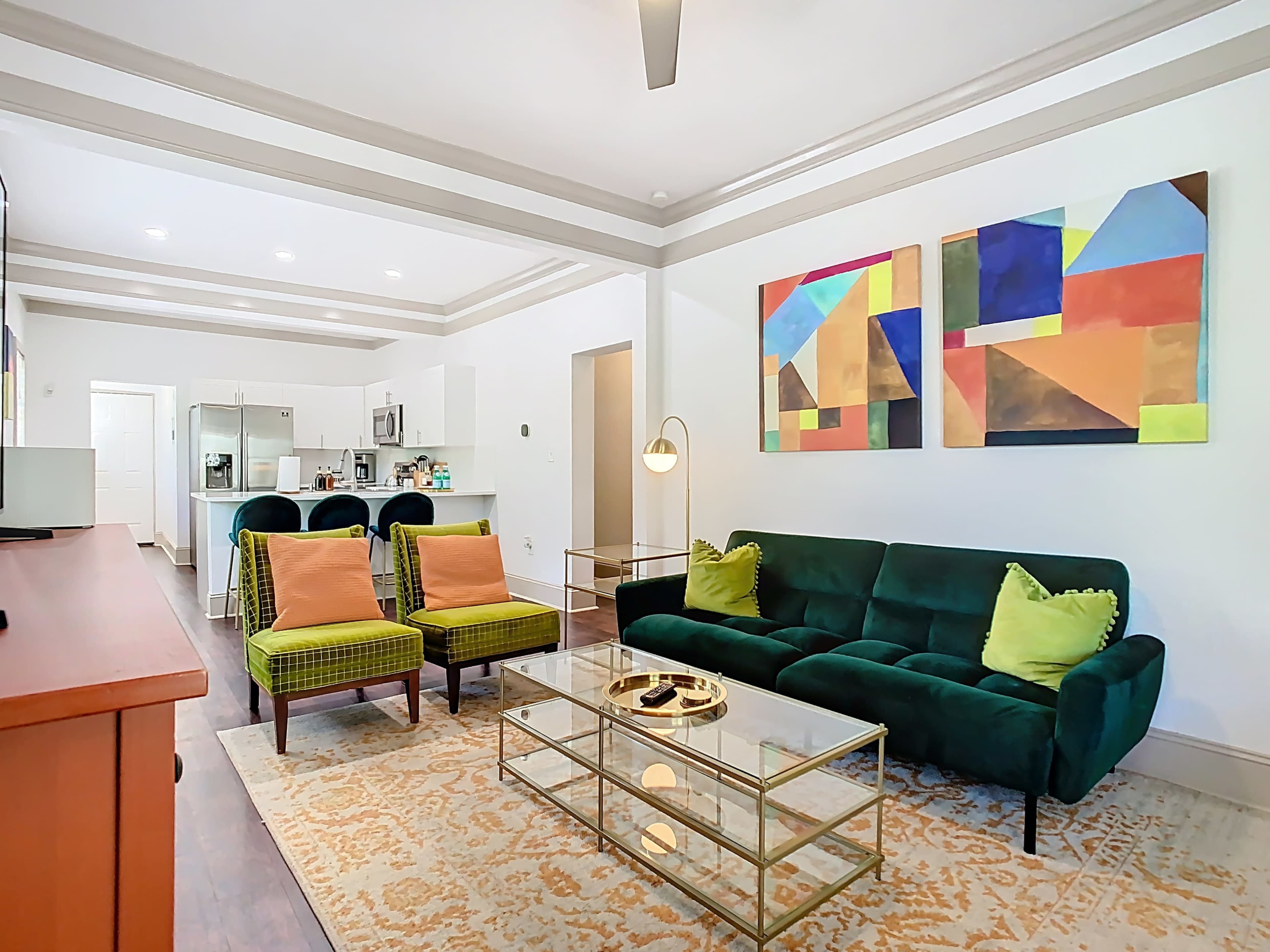 The image shows a modern living room featuring a green sofa, two patterned chairs, a glass coffee table, and an abstract painting on the wall.