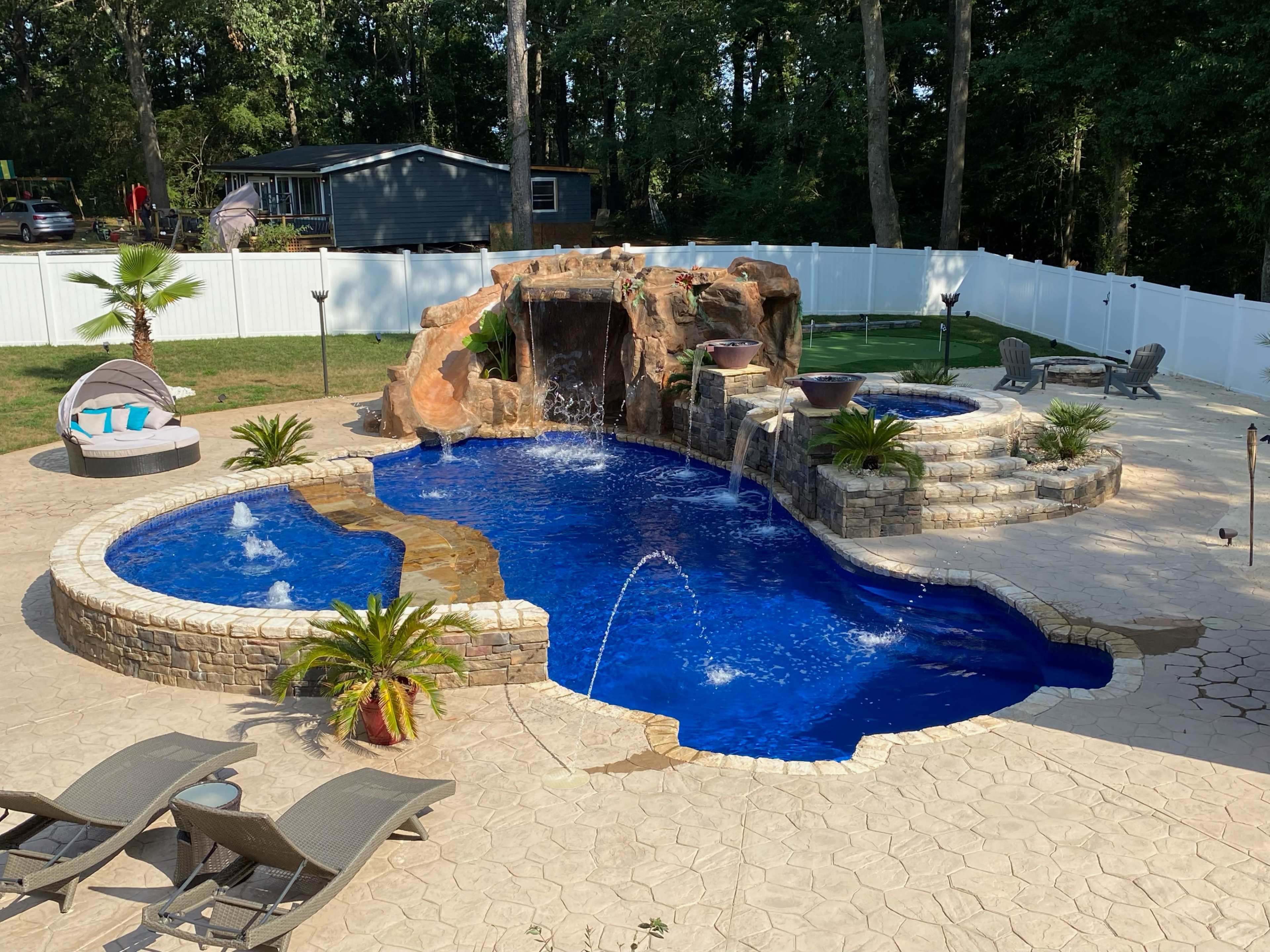 A landscaped backyard featuring a blue swimming pool with a rock waterfall, surrounded by stone coping, tropical plants, and poolside furniture.