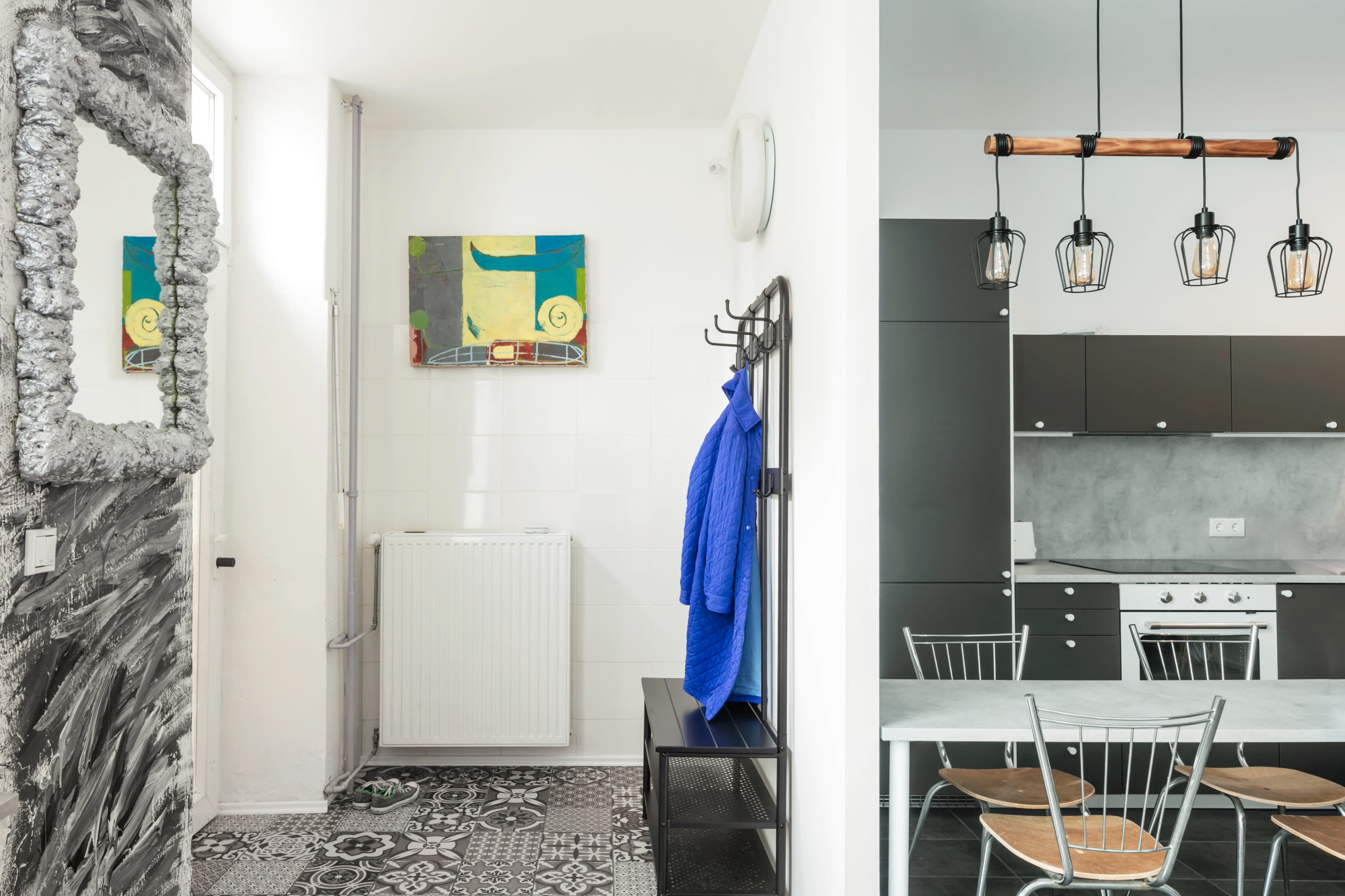 The image shows a modern entryway with a patterned floor, a mirror, a coat rack with a blue jacket, and a view into a kitchen area featuring gray cabinets and a dining table with metal chairs.