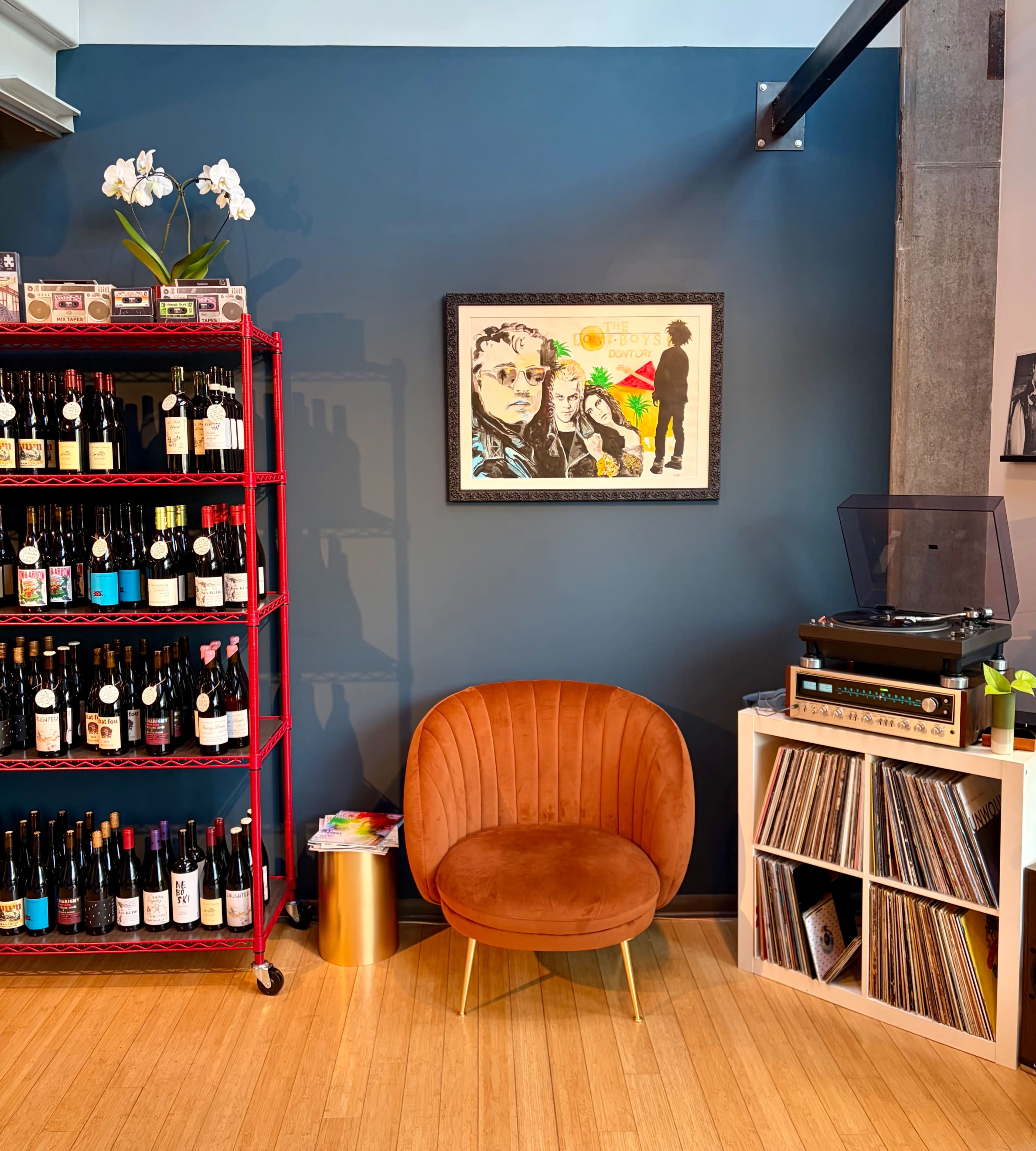 The image features a cozy corner of a room with a vinyl record player, a collection of wine bottles on a metal rack, and a framed artwork on the wall above a curved orange chair.