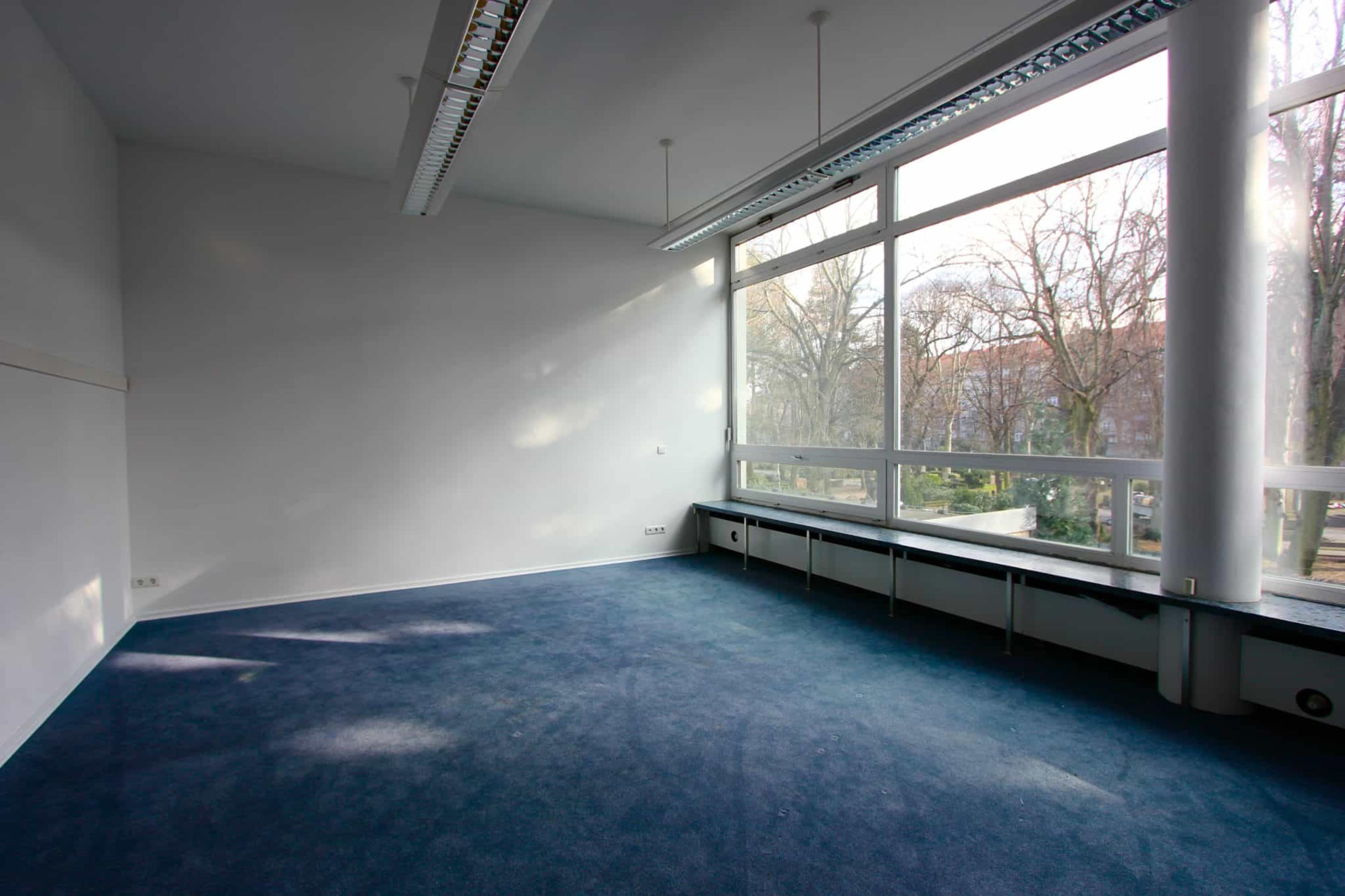 The image shows a sparsely furnished office space with blue carpeting and large windows allowing natural light to enter.