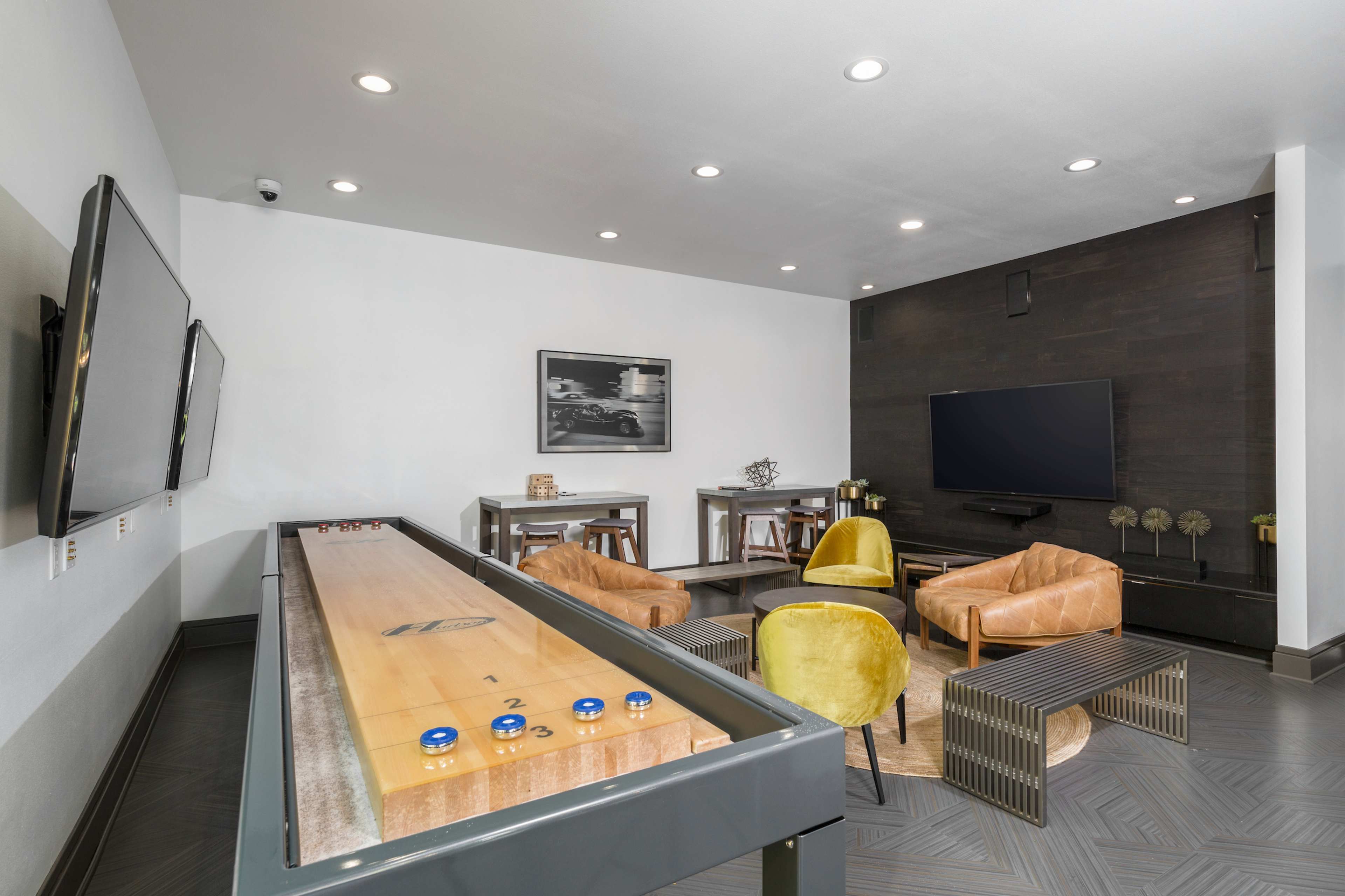 A modern leisure room featuring a shuffleboard table, yellow armchairs, and two large flat-screen TVs mounted on the walls.