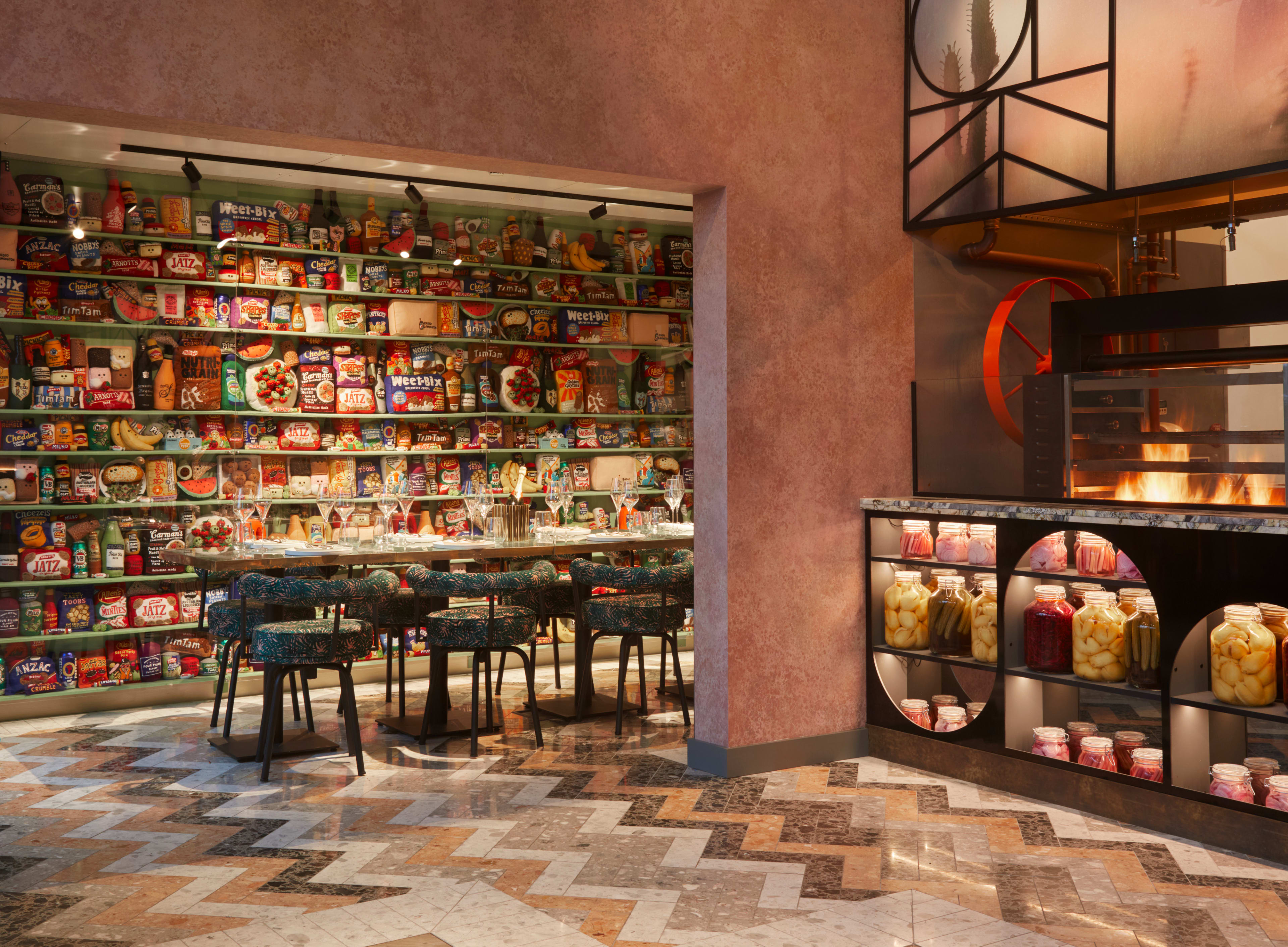 A brightly designed restaurant features a wall of colorful packaged foods and a cooking area with a visible oven, while stylish seating is arranged around a central table.