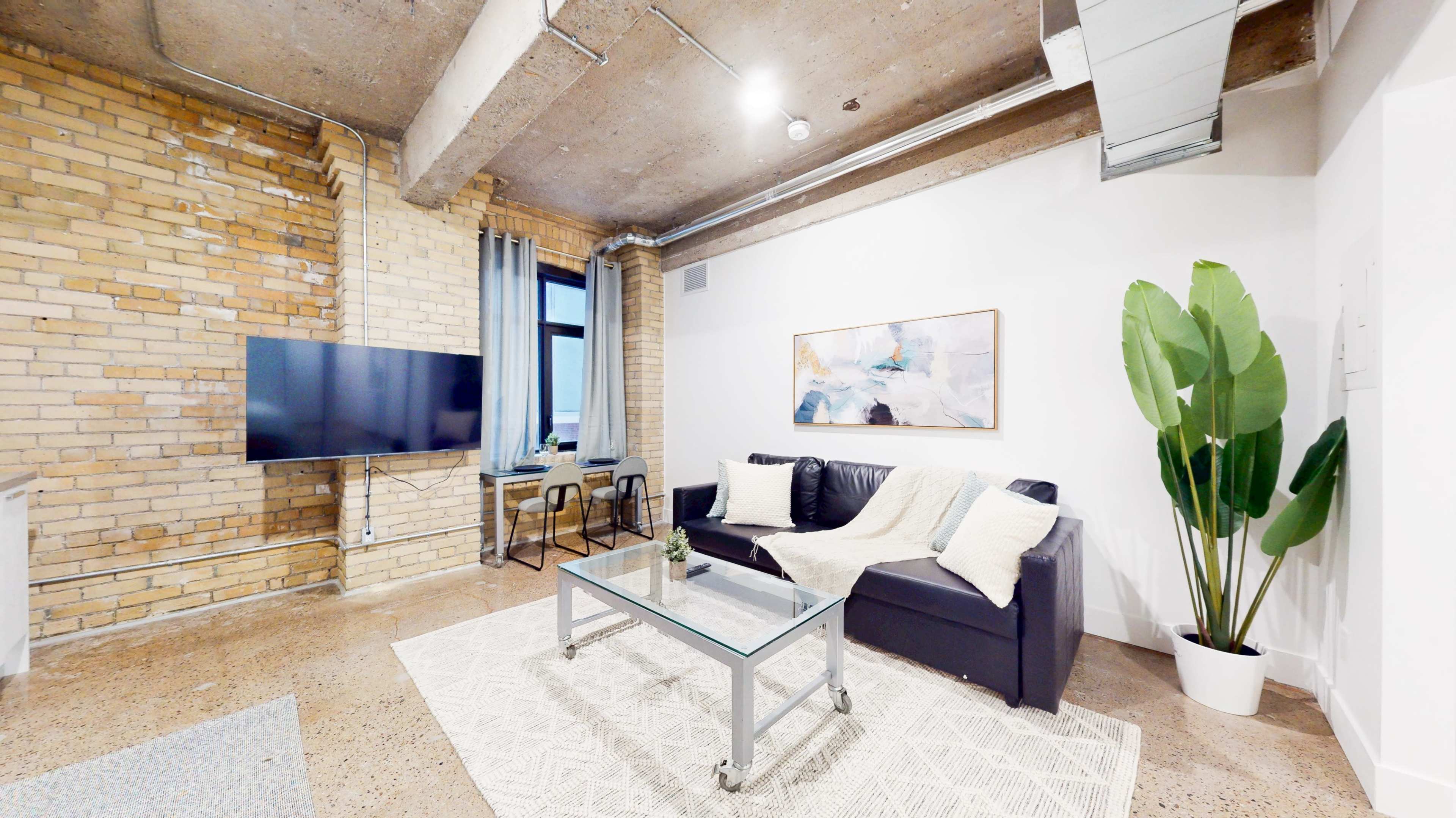 The image shows a modern living room with exposed brick walls, a black sofa, a glass coffee table, and a large potted plant in the corner.