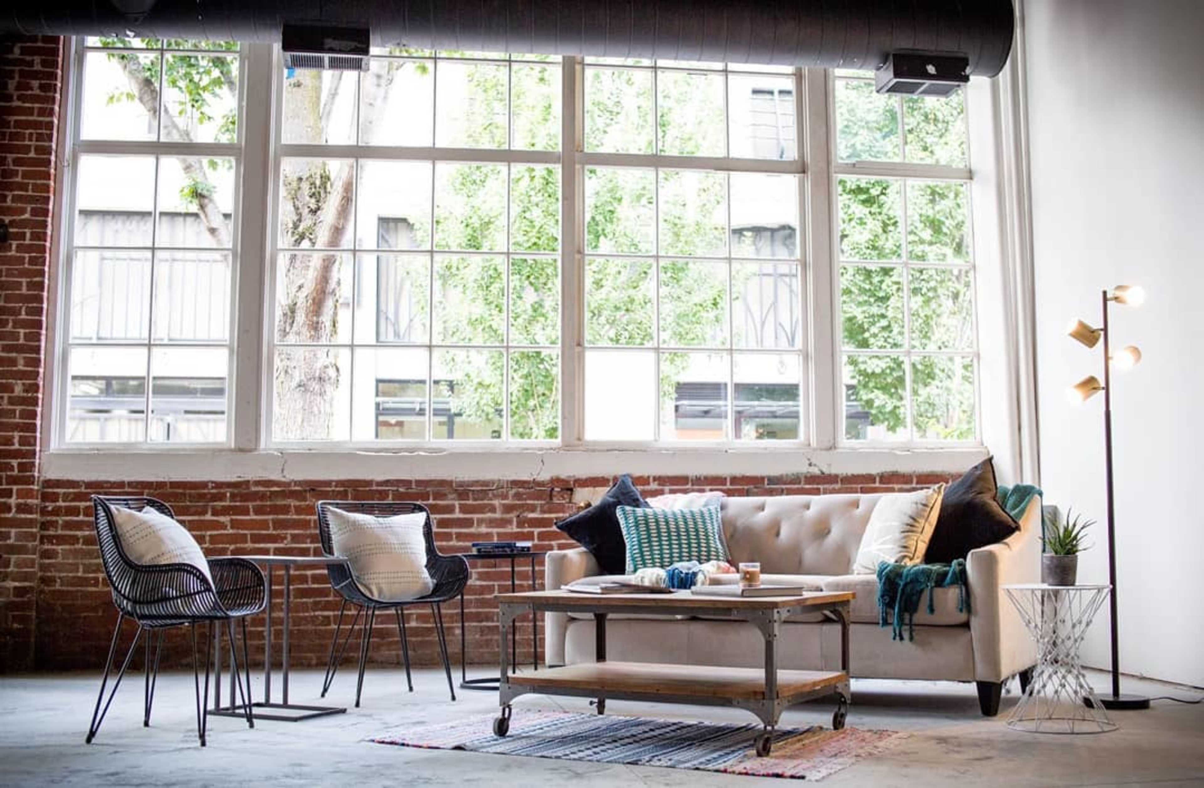 The scene features a cozy living room with a beige sofa, two wire chairs, a coffee table, and large windows allowing natural light to enter.