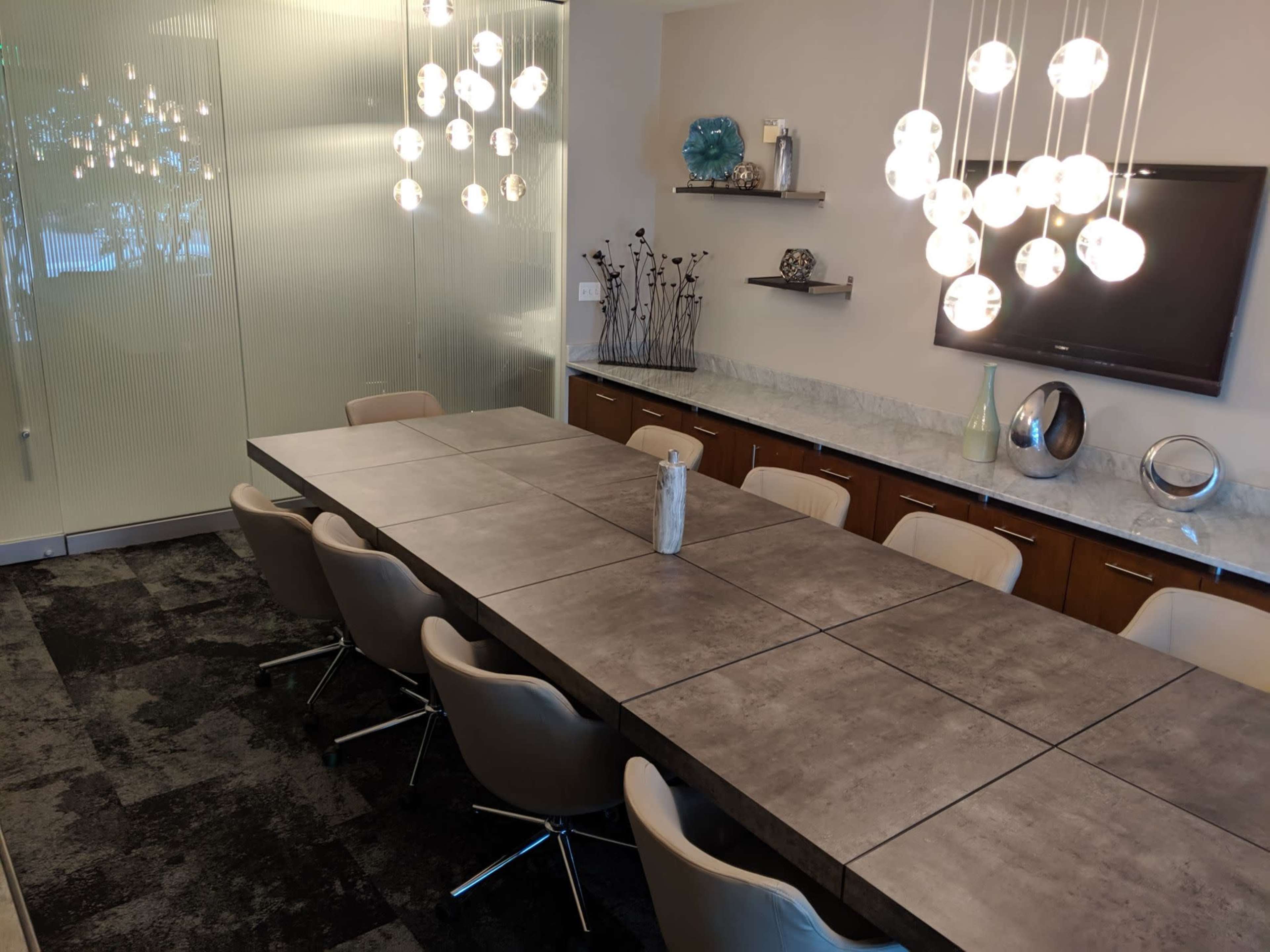 A modern conference room features a long, rectangular table surrounded by upholstered chairs, with pendant lights hanging above and a glass panel wall.