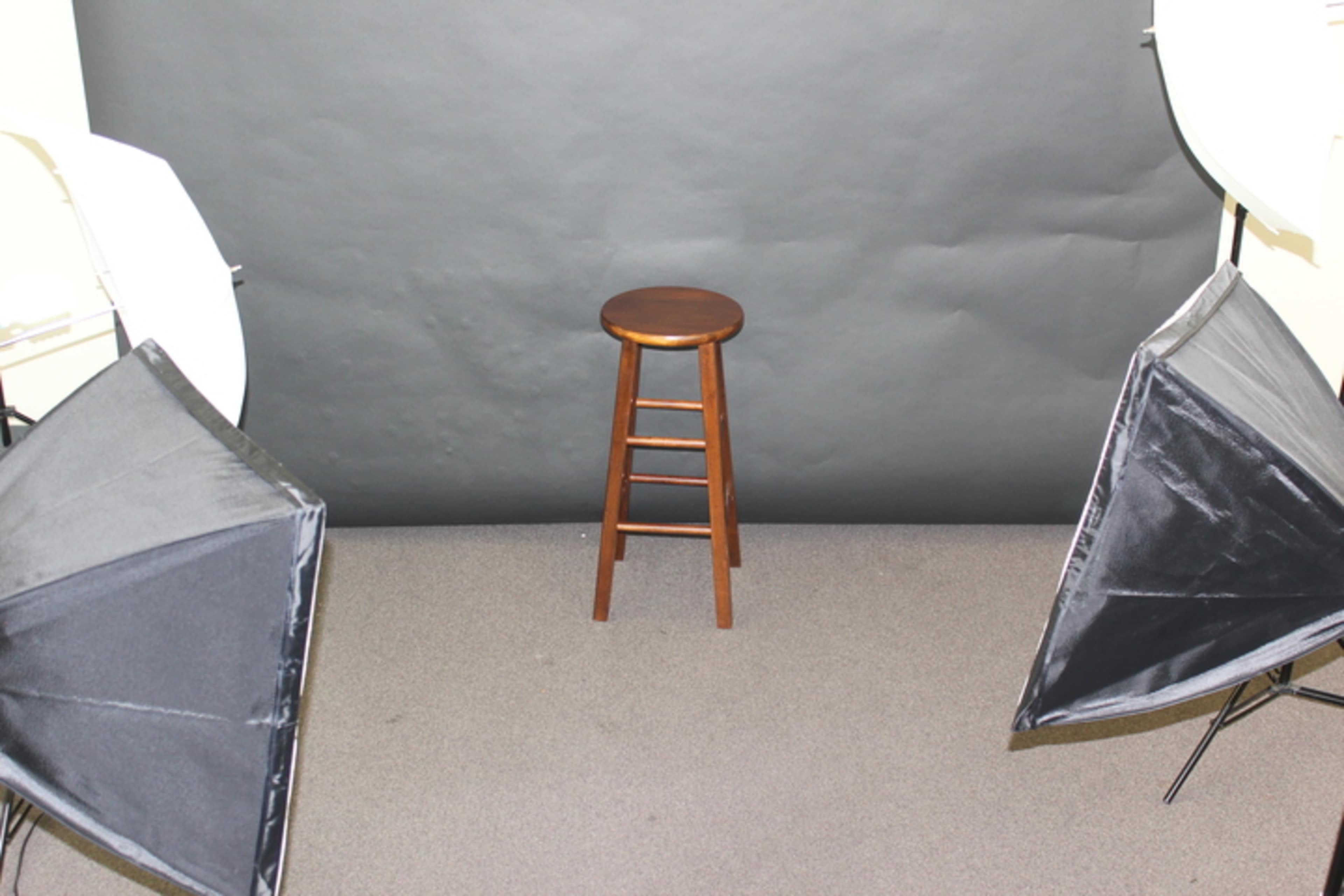 A wooden stool is positioned in front of a gray backdrop, surrounded by two softbox lights.