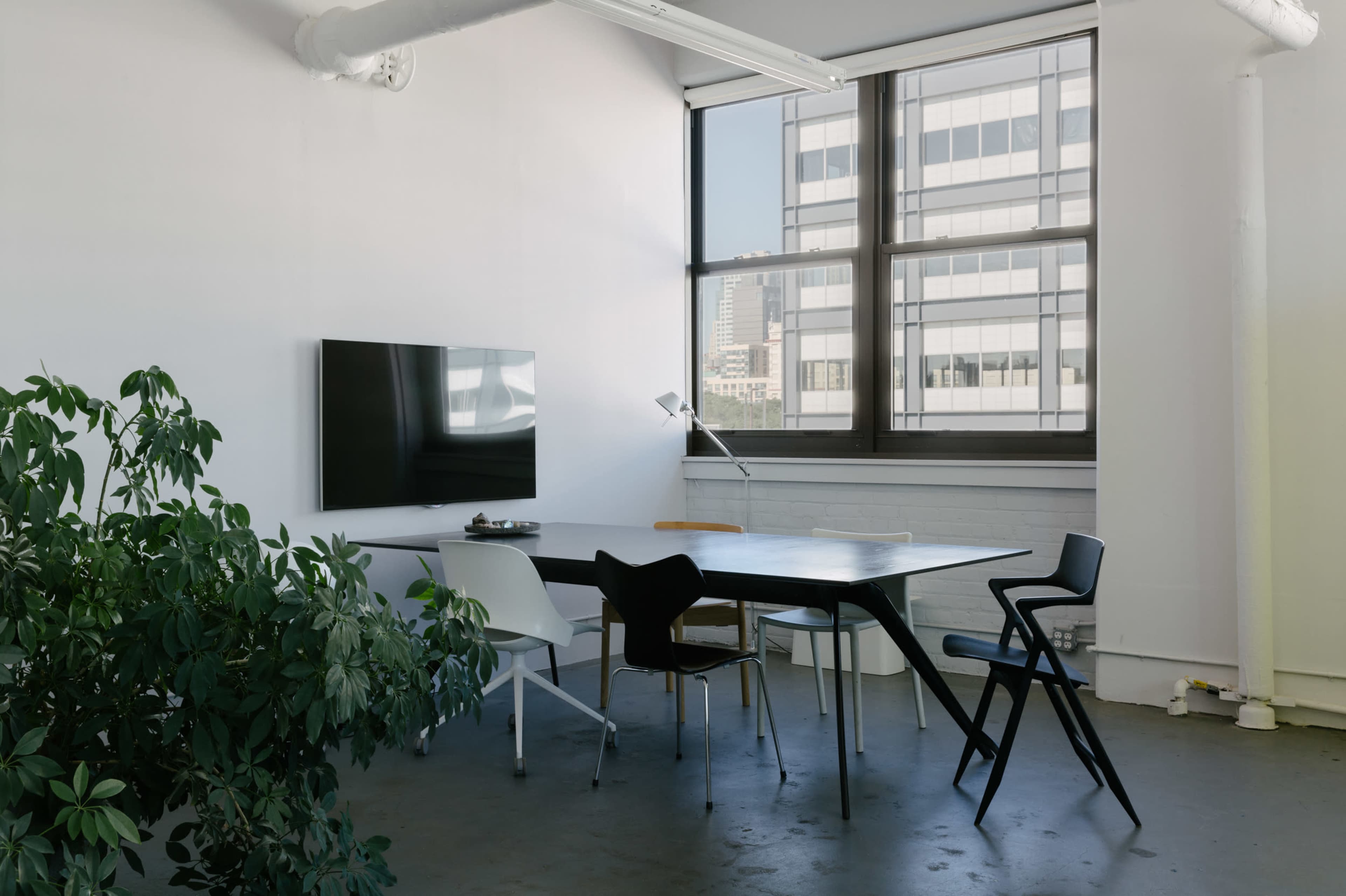 A minimalist office space features a large table surrounded by chairs, with a television mounted on the wall and a plant in the corner.