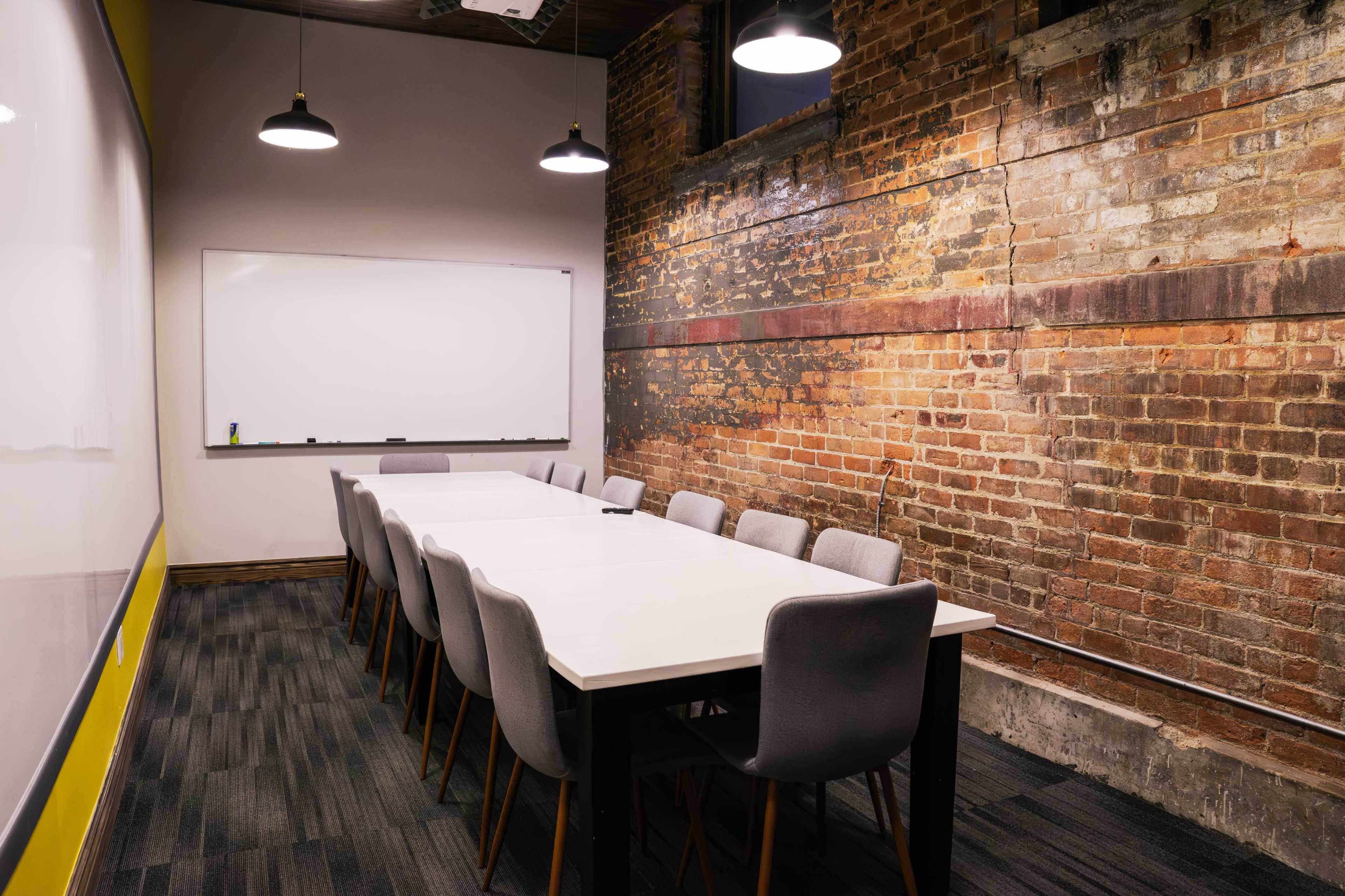 A conference room features a long white table surrounded by gray chairs, with exposed brick walls and a whiteboard on the front wall.