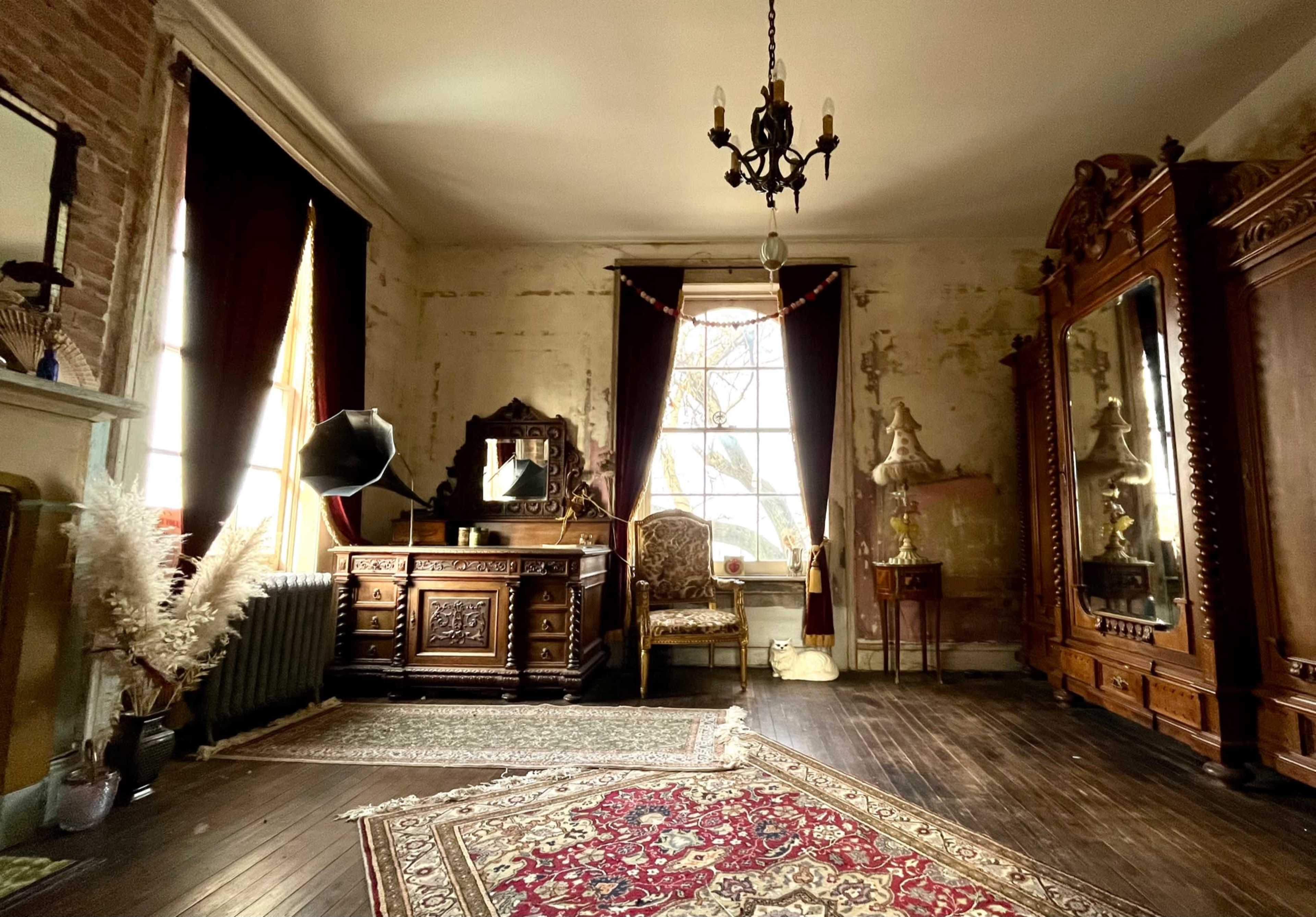 An ornate room featuring a large wooden dresser, a vintage mirror, and a patterned area rug, illuminated by natural light through tall windows.