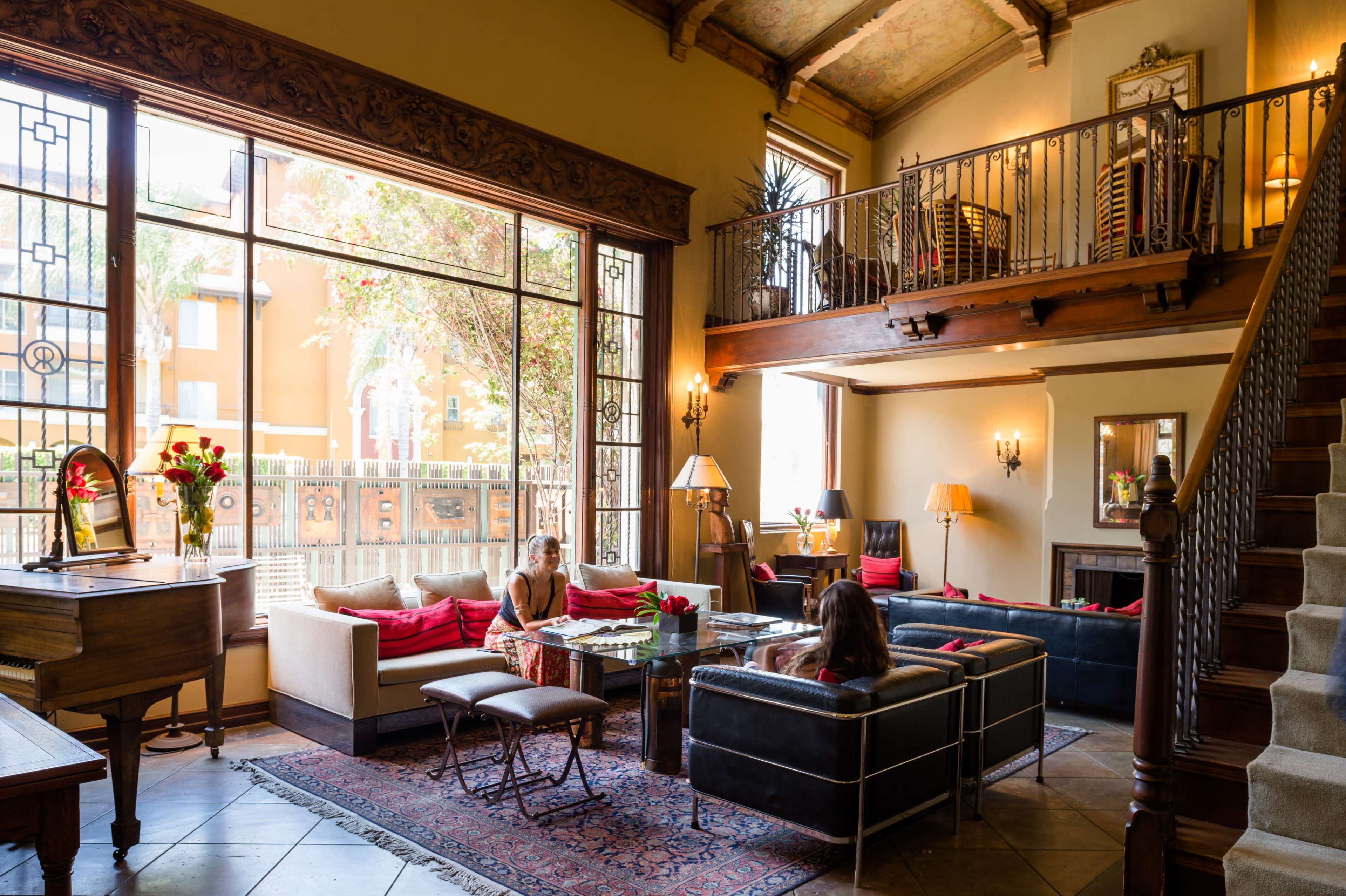 The image shows a spacious living room with large windows, a staircase, and various seating arrangements around a central coffee table.