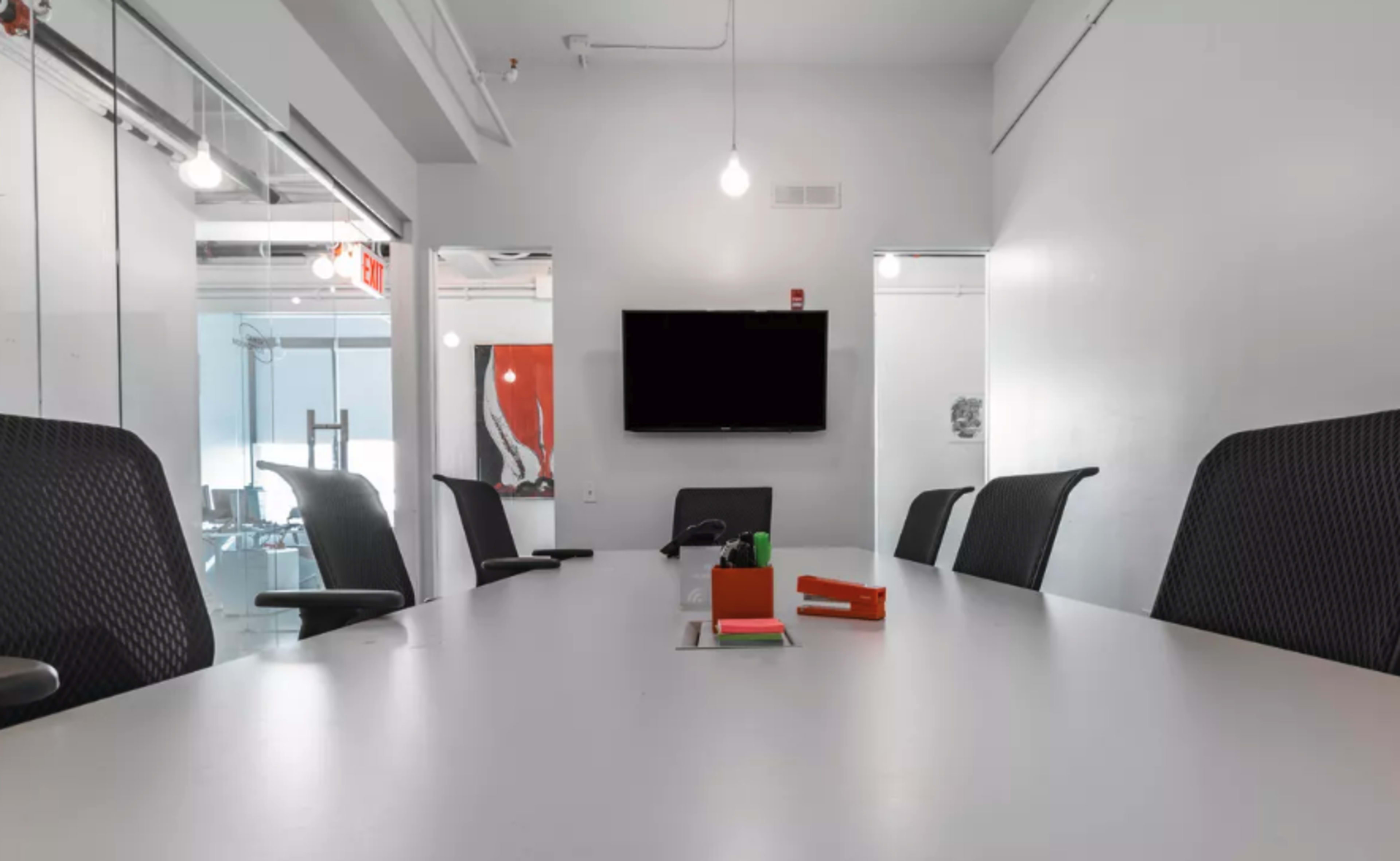 A conference room features a long white table, black mesh chairs, and a wall-mounted television.