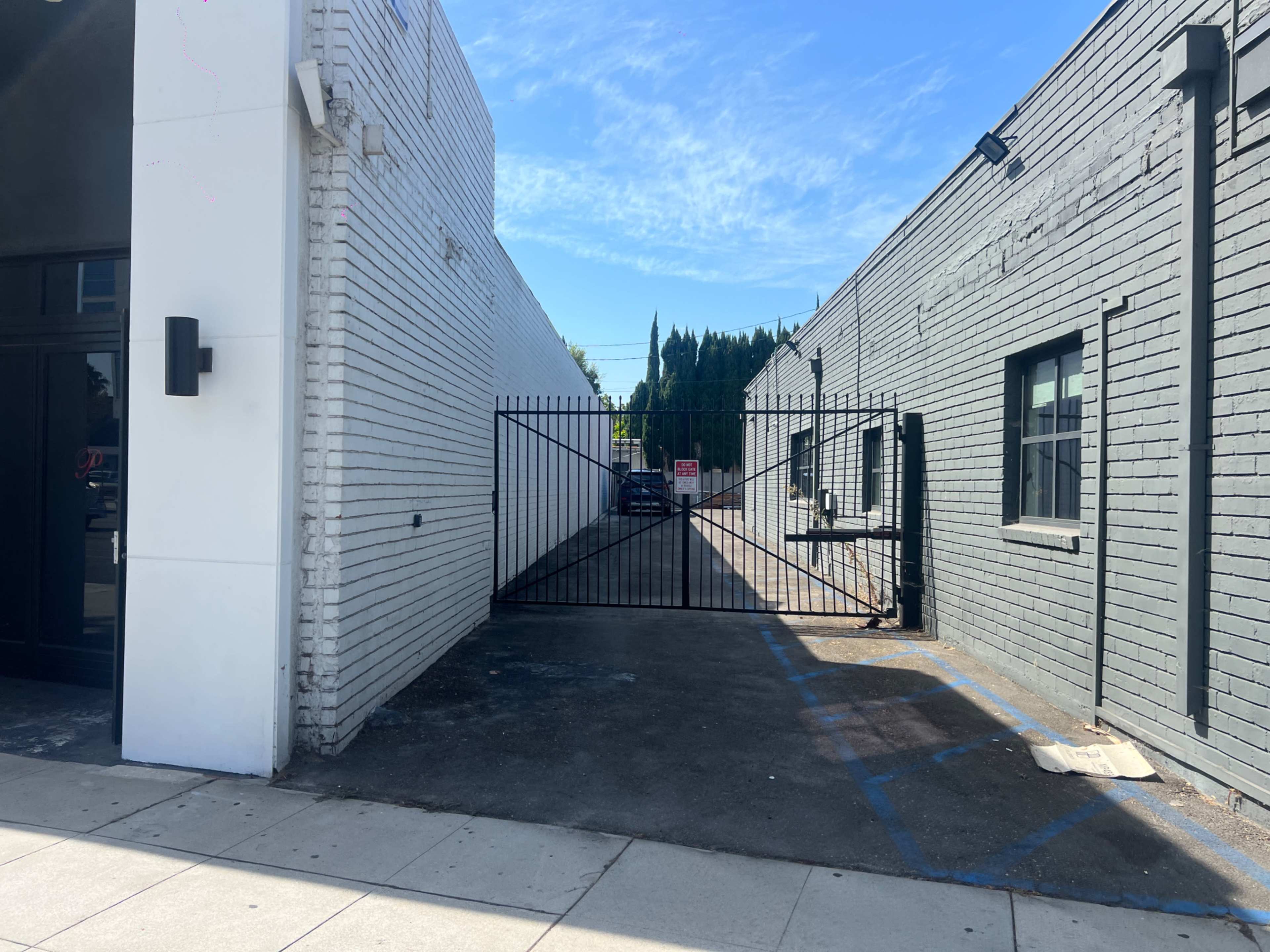 A narrow alleyway with a gated entrance is flanked by two buildings, one white and the other gray, under a clear blue sky.