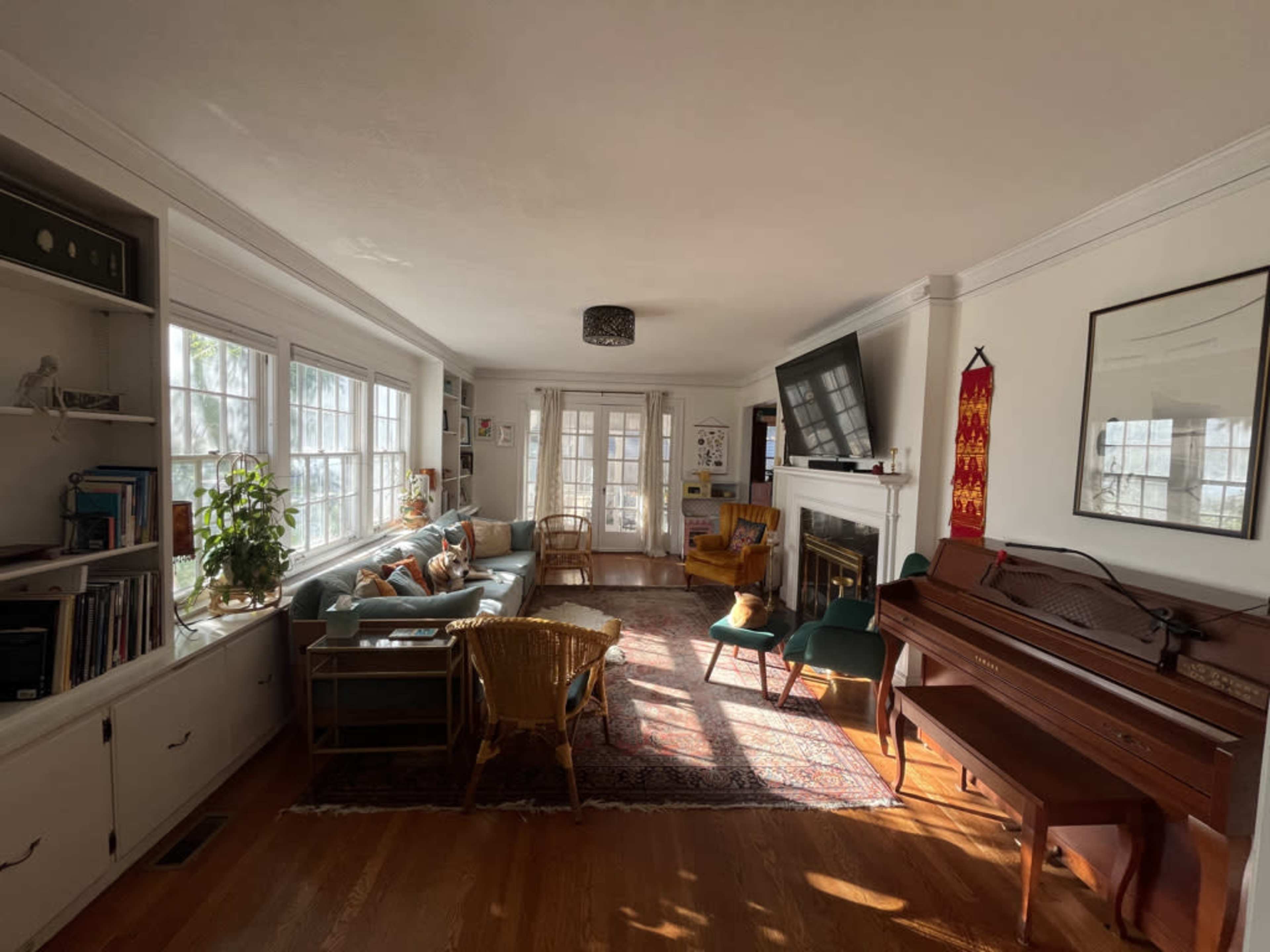 The image shows a bright living room with large windows, a seating area featuring a couch and chairs, a piano, and a fireplace.