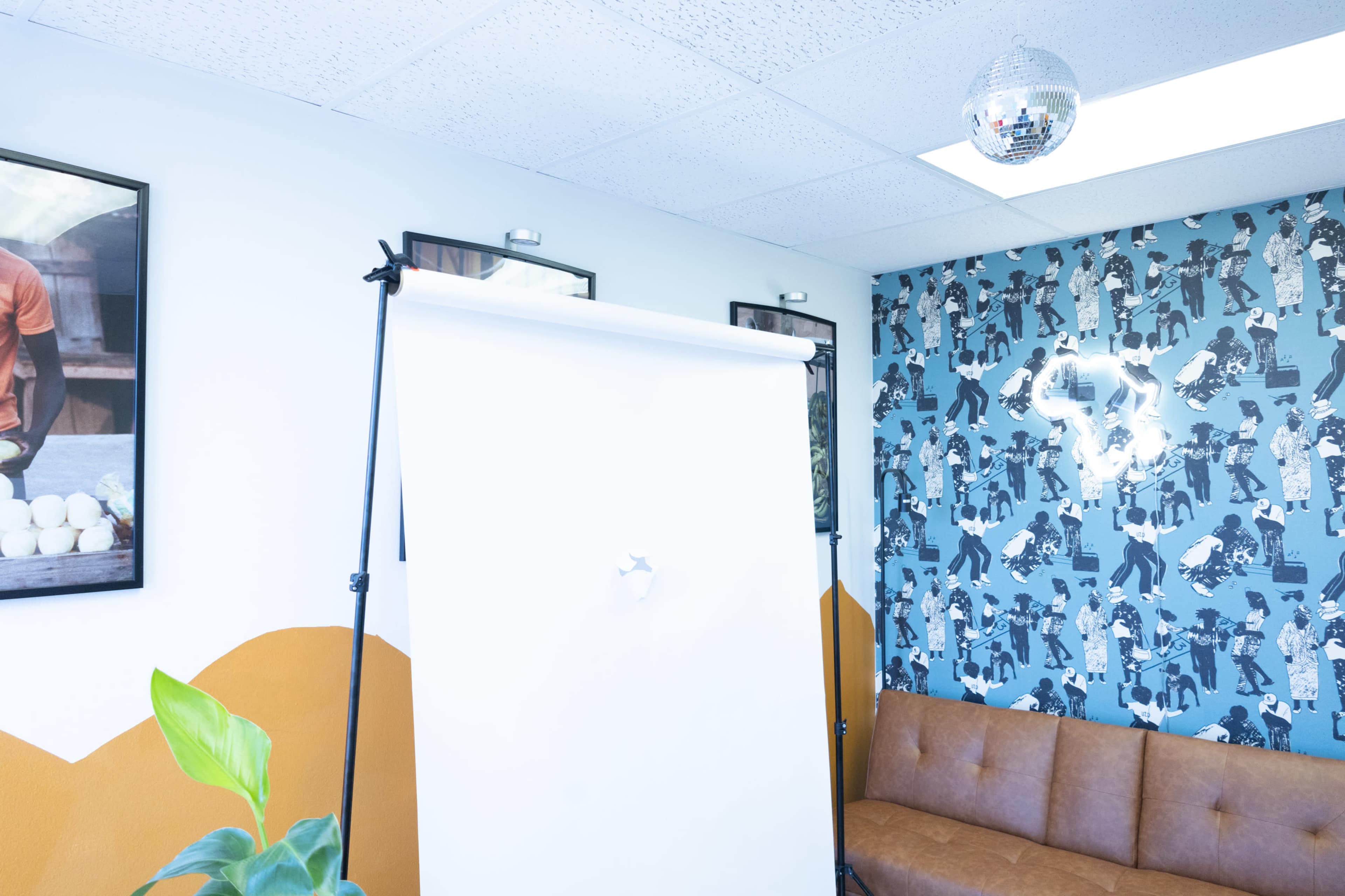 A small room with a large white backdrop on a stand, a brown couch, and patterned wallpaper depicting various figures, alongside framed pictures on the walls and a disco ball hanging from the ceiling.
