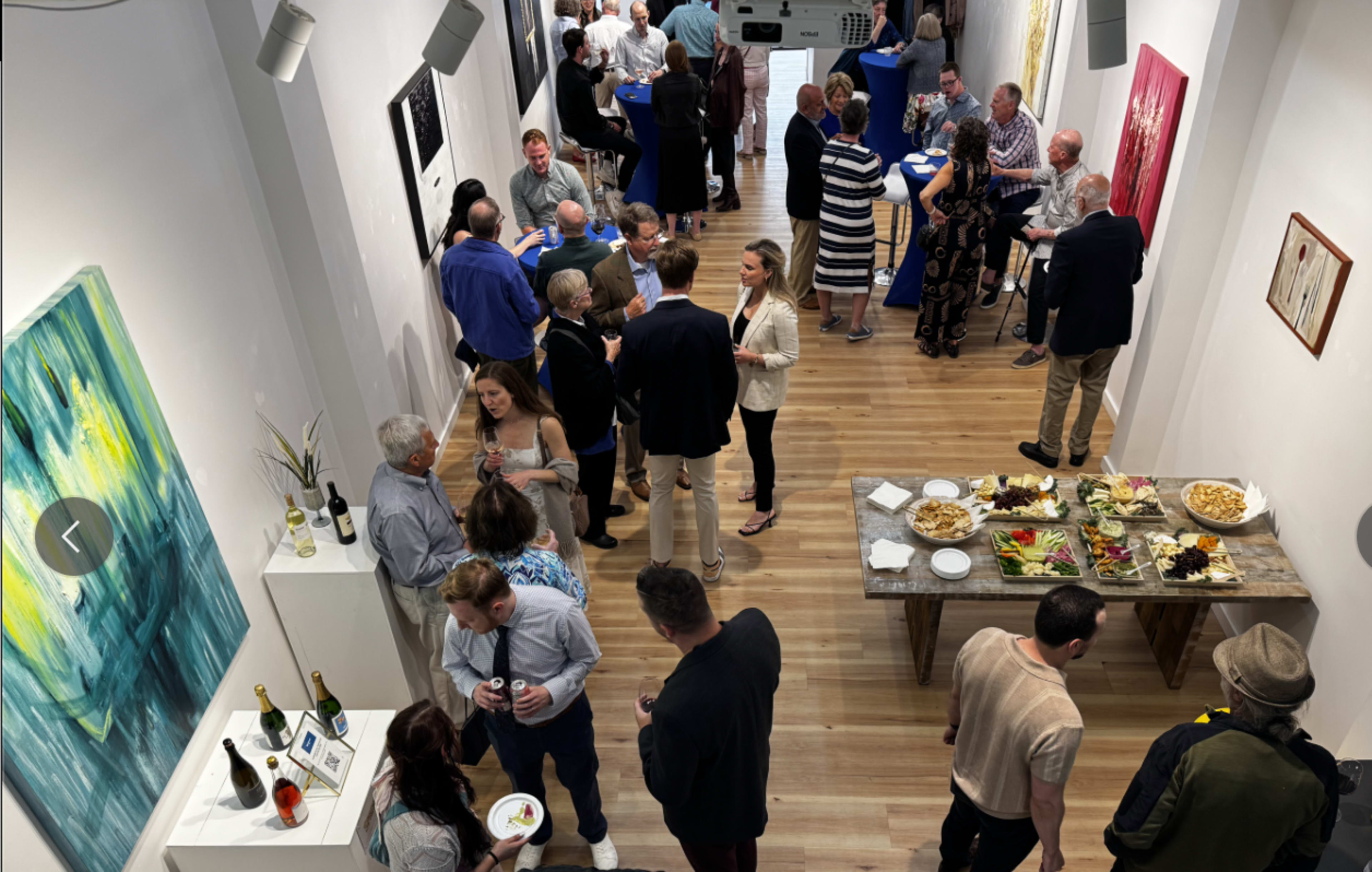 The image shows a crowded art gallery event with people mingling and interacting amidst various artworks on the walls and a table filled with food and drinks.