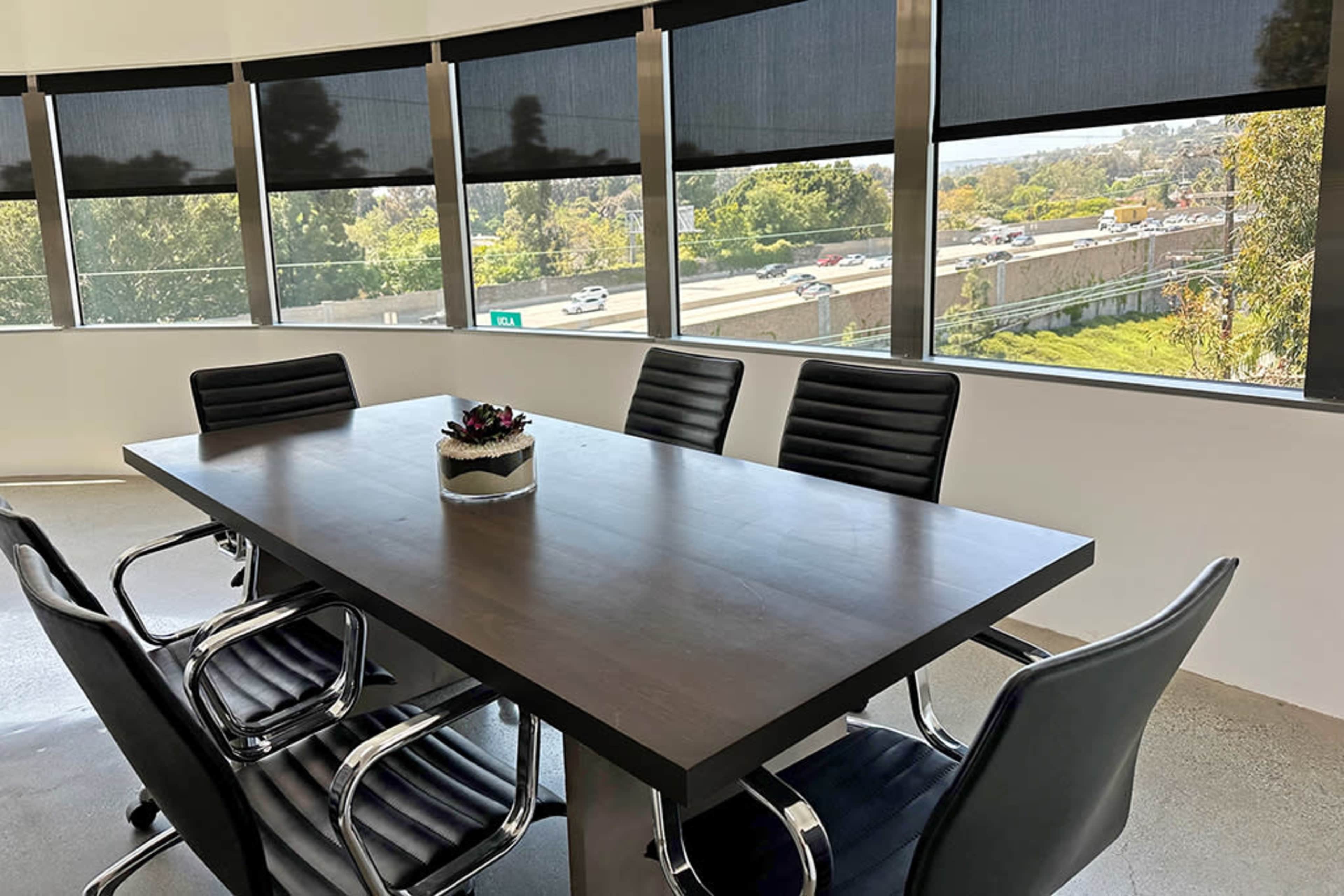 A modern conference room features a large table surrounded by black chairs, with large windows showcasing a view of a highway and greenery outside.