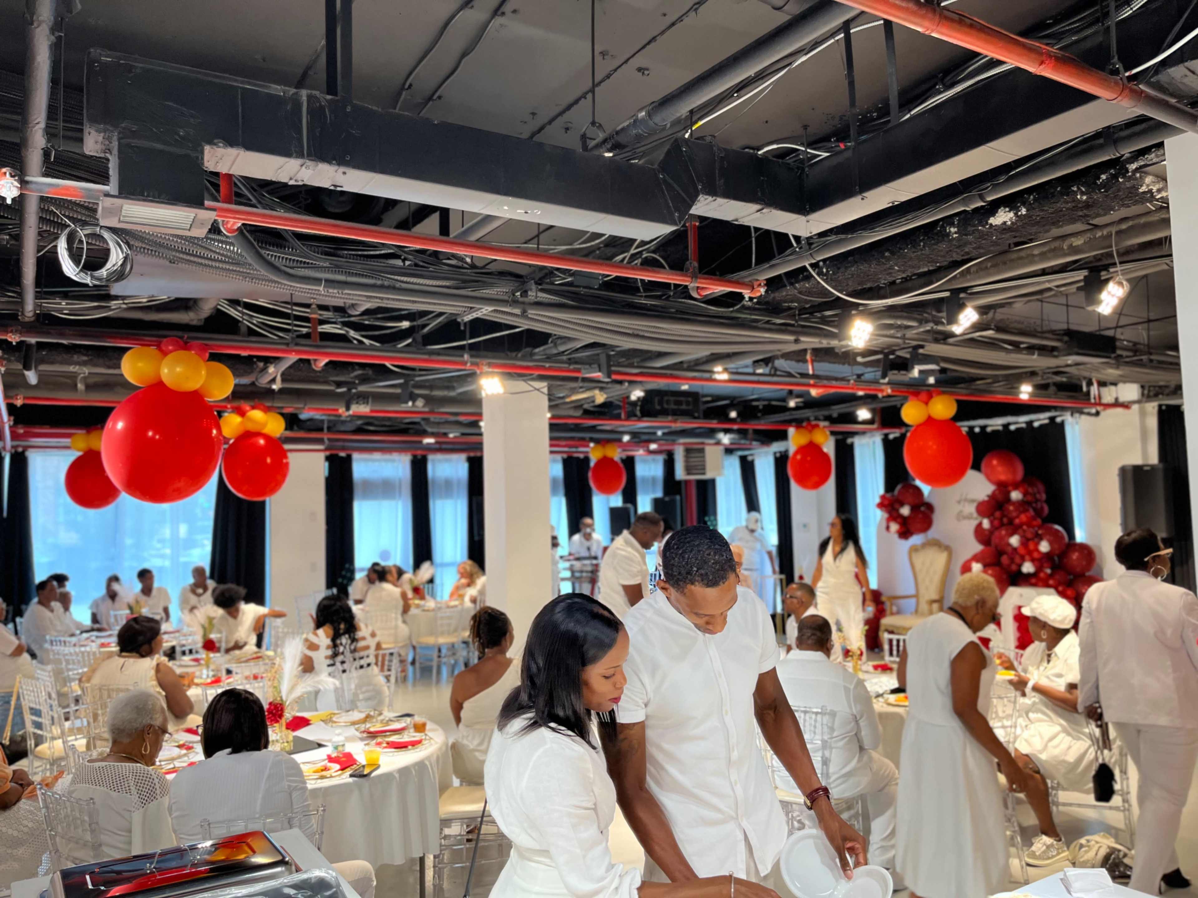 A dining area is set up for an event, featuring guests in white attire seated at tables adorned with red and yellow decorations.