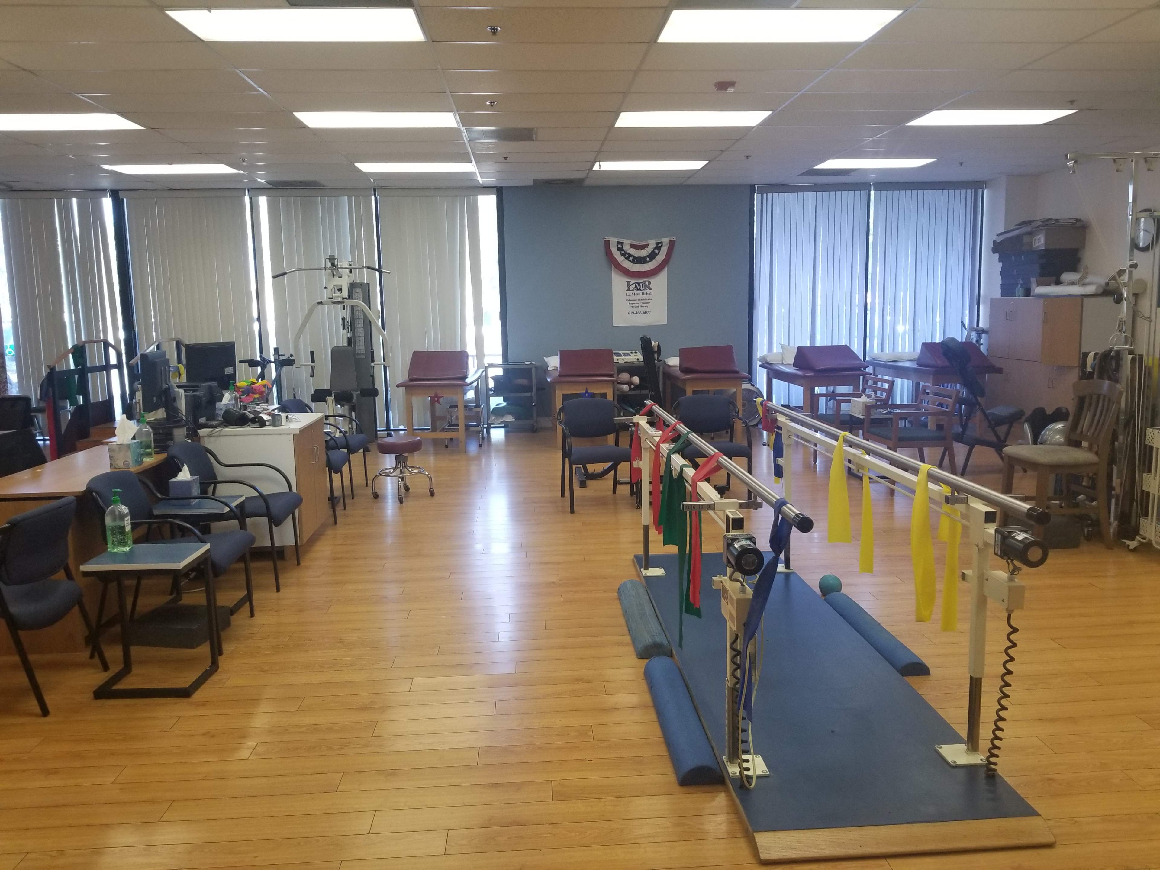 A therapy room with exercise equipment, including a pulley system and various therapy mats, surrounded by chairs and workstations.