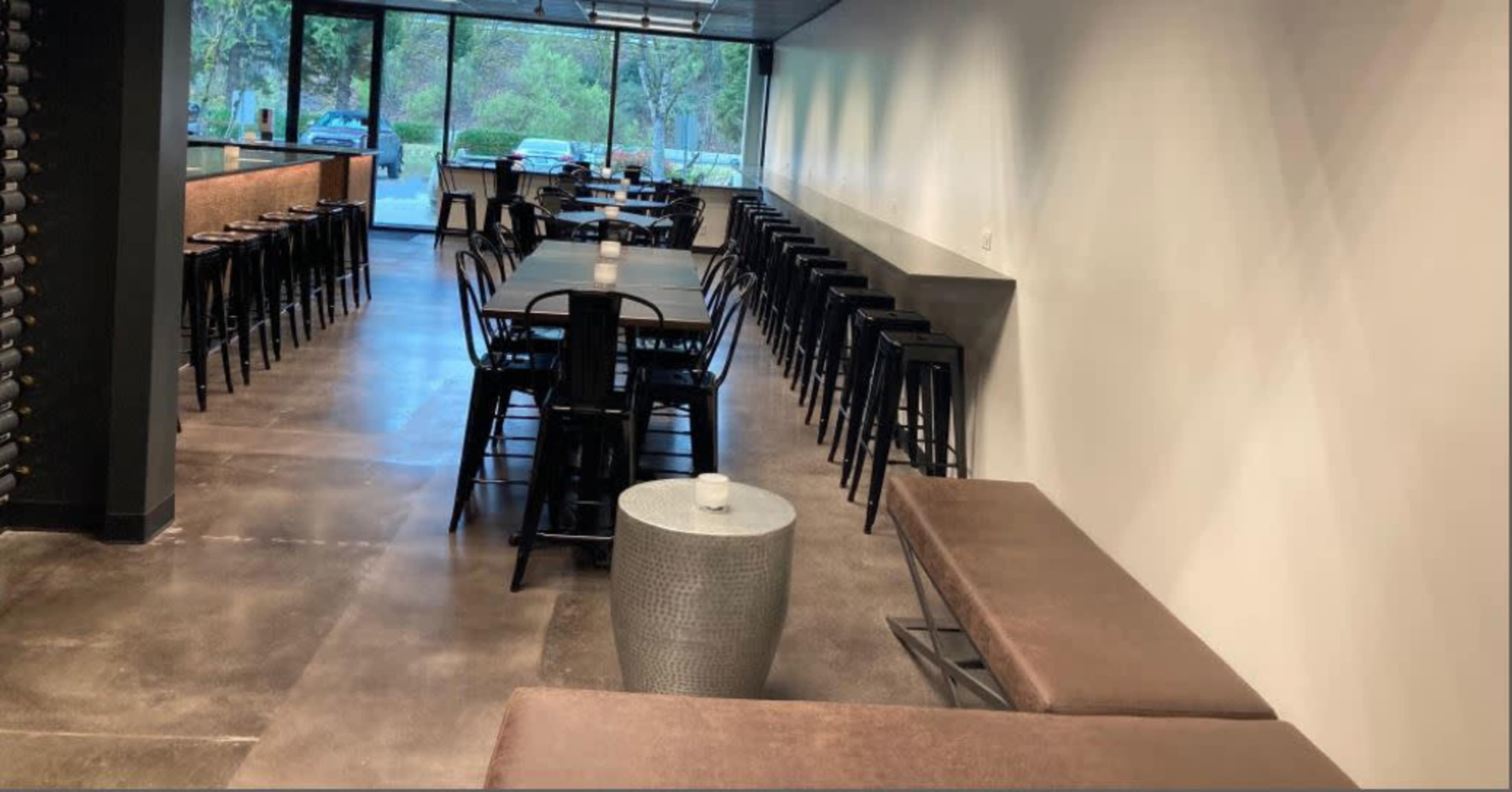 A modern dining area with tables lined up along the walls and a row of chairs facing a large window that overlooks an outdoor space.