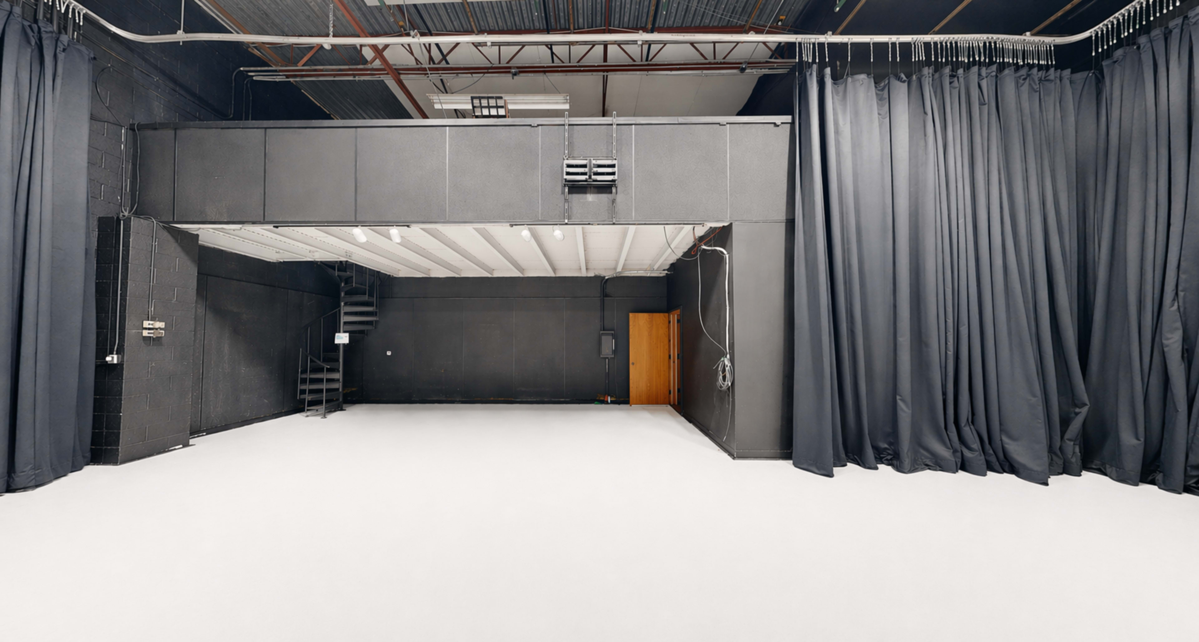 The image shows a spacious black-walled studio with a raised area and black curtains on one side.