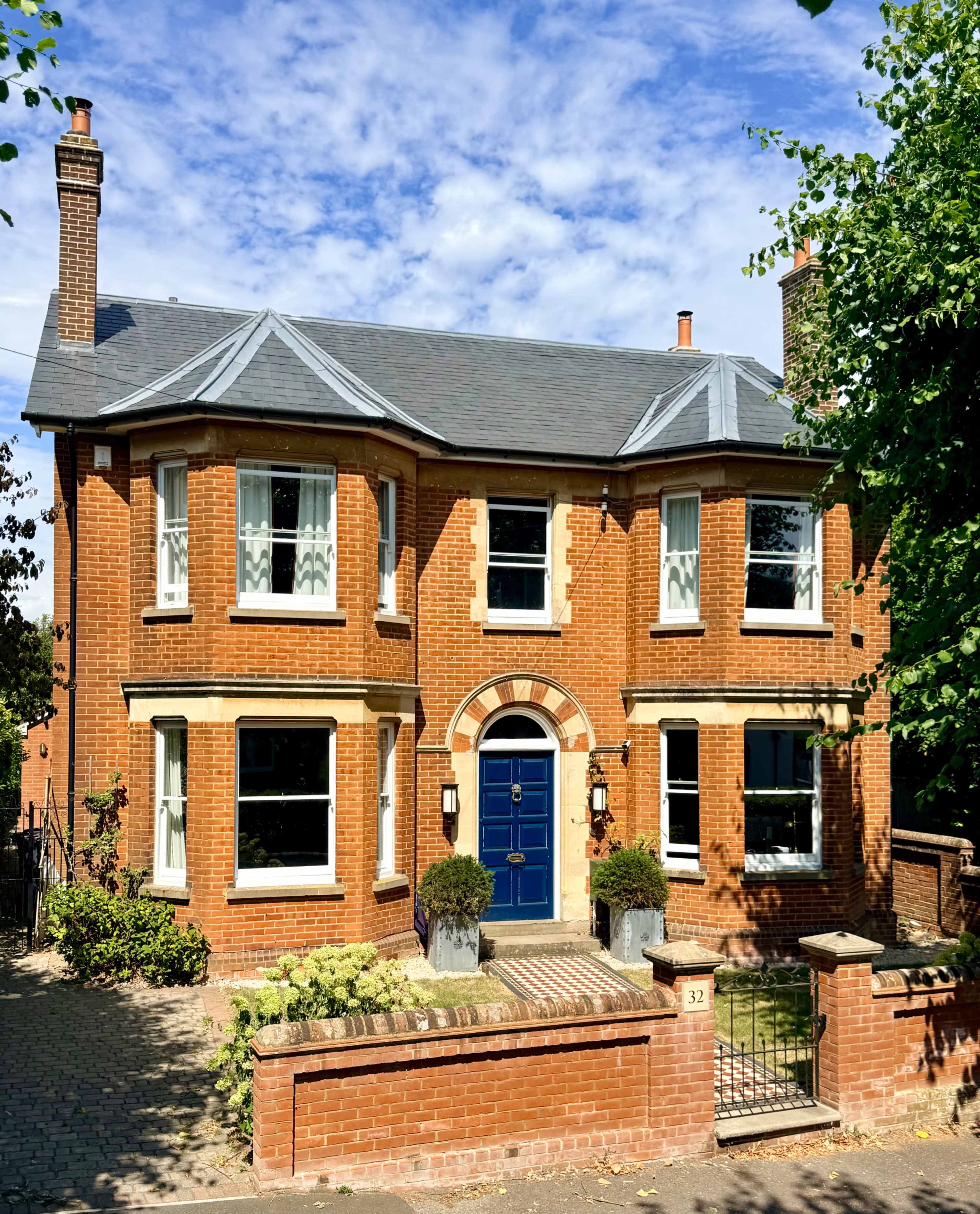The image shows a two-story brick house with a gabled roof and large windows, fronted by a garden and a brick wall.