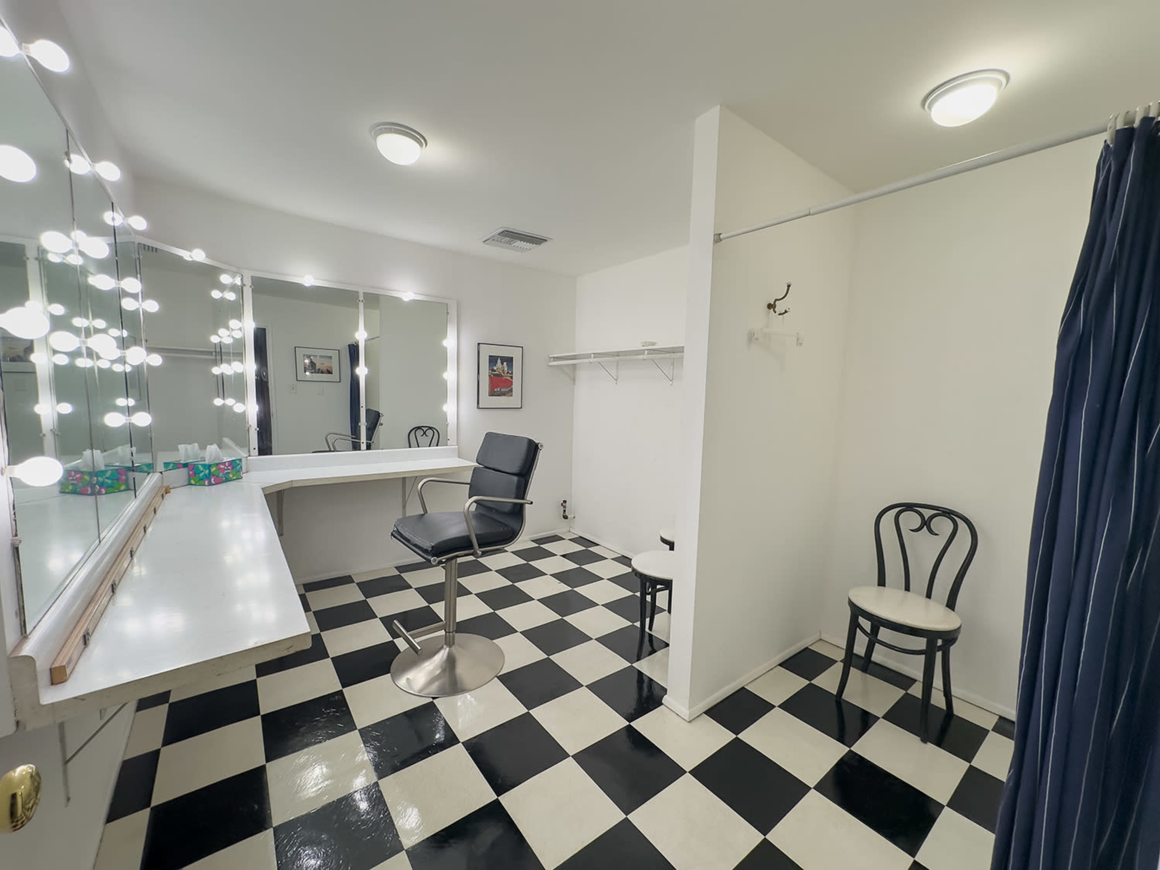 A brightly lit dressing room with a mirrored vanity, a black and white checked floor, and minimal furnishings including two chairs and a clothing rack.