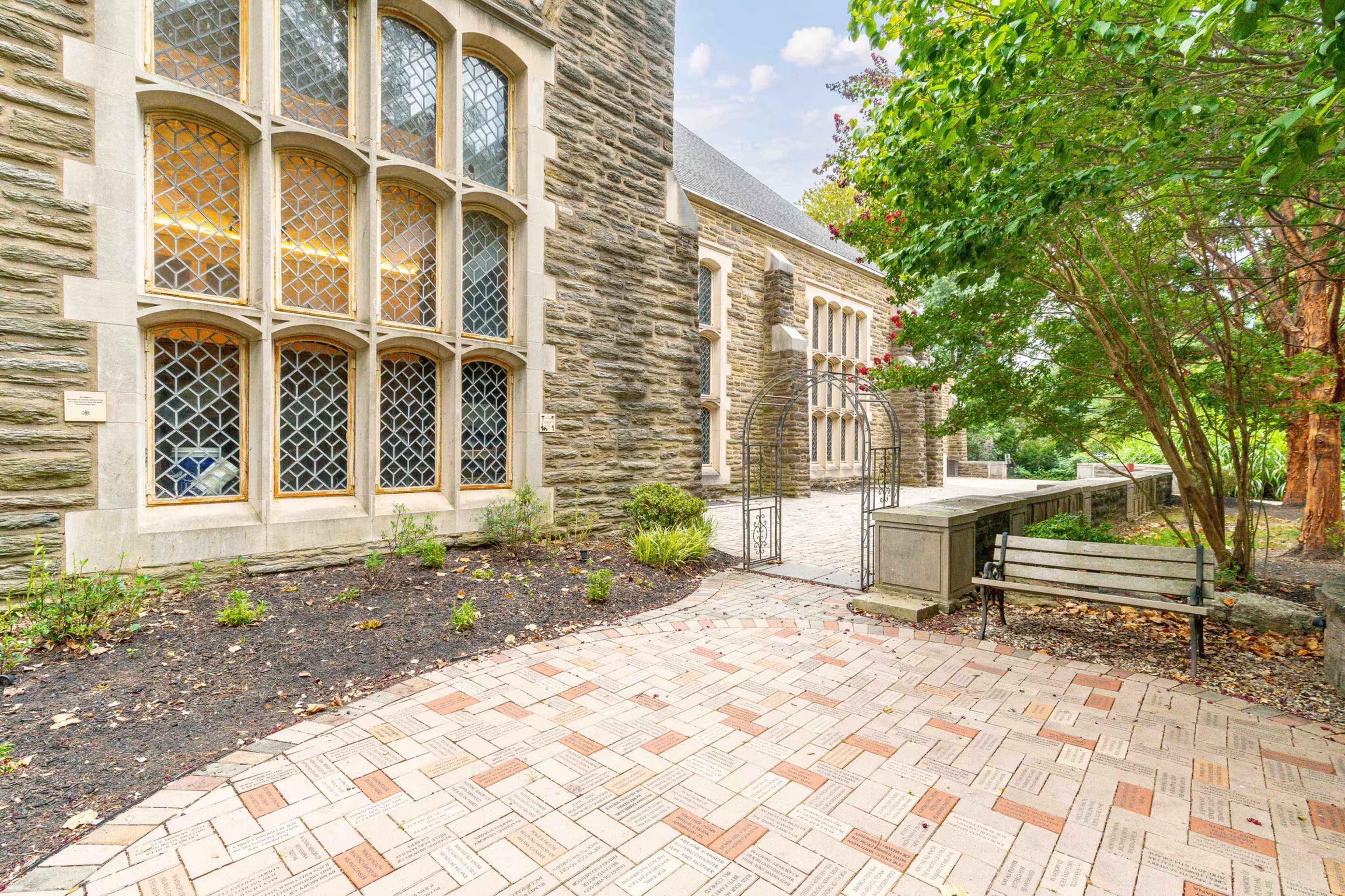 The image shows a stone building with large, arched, leaded glass windows beside a brick pathway and a wooden bench, framed by green trees and shrubs.