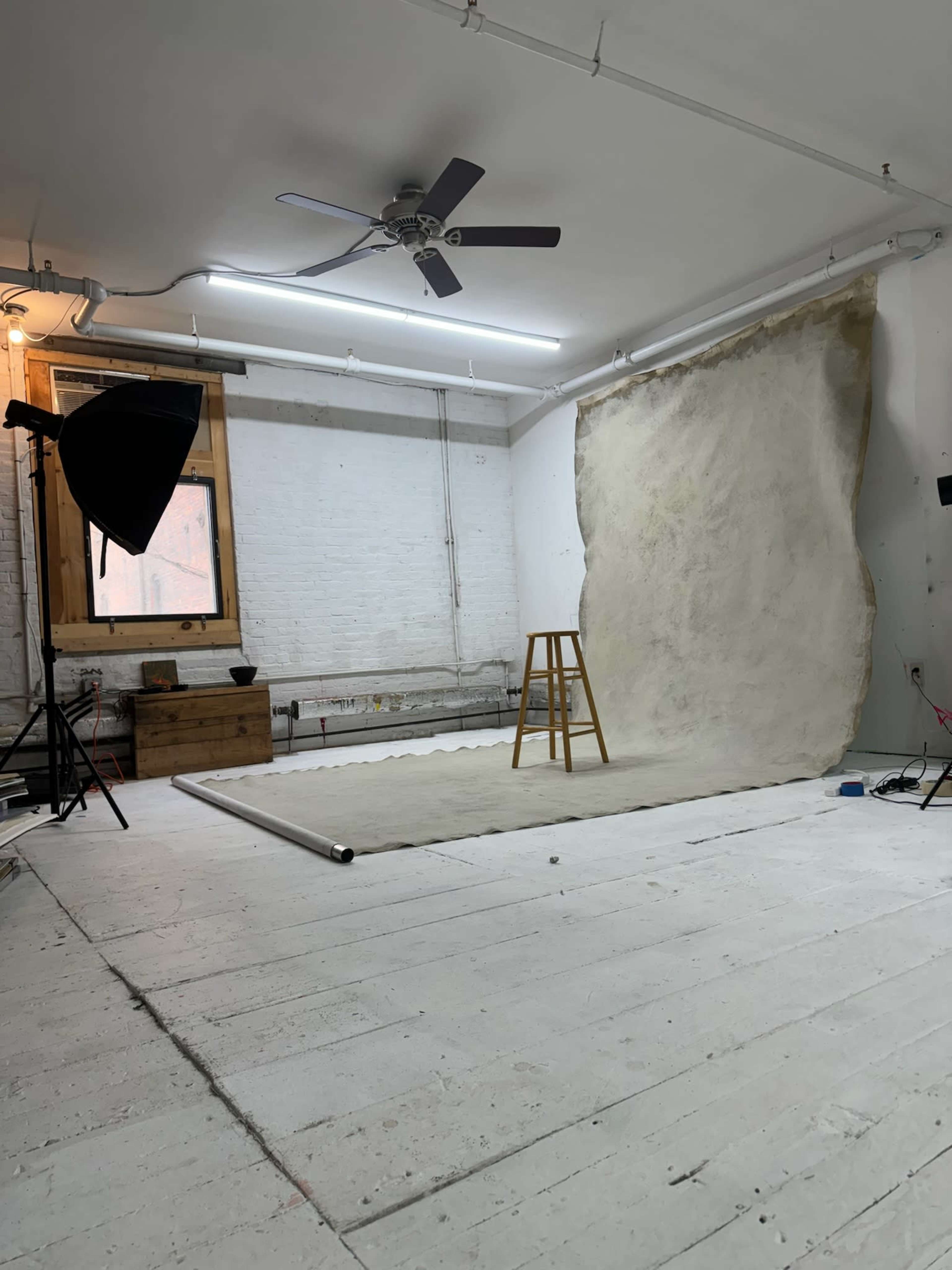 A photography studio is set up with a wooden stool, a backdrop partially rolled out, and lighting equipment arranged around a whitewashed room.