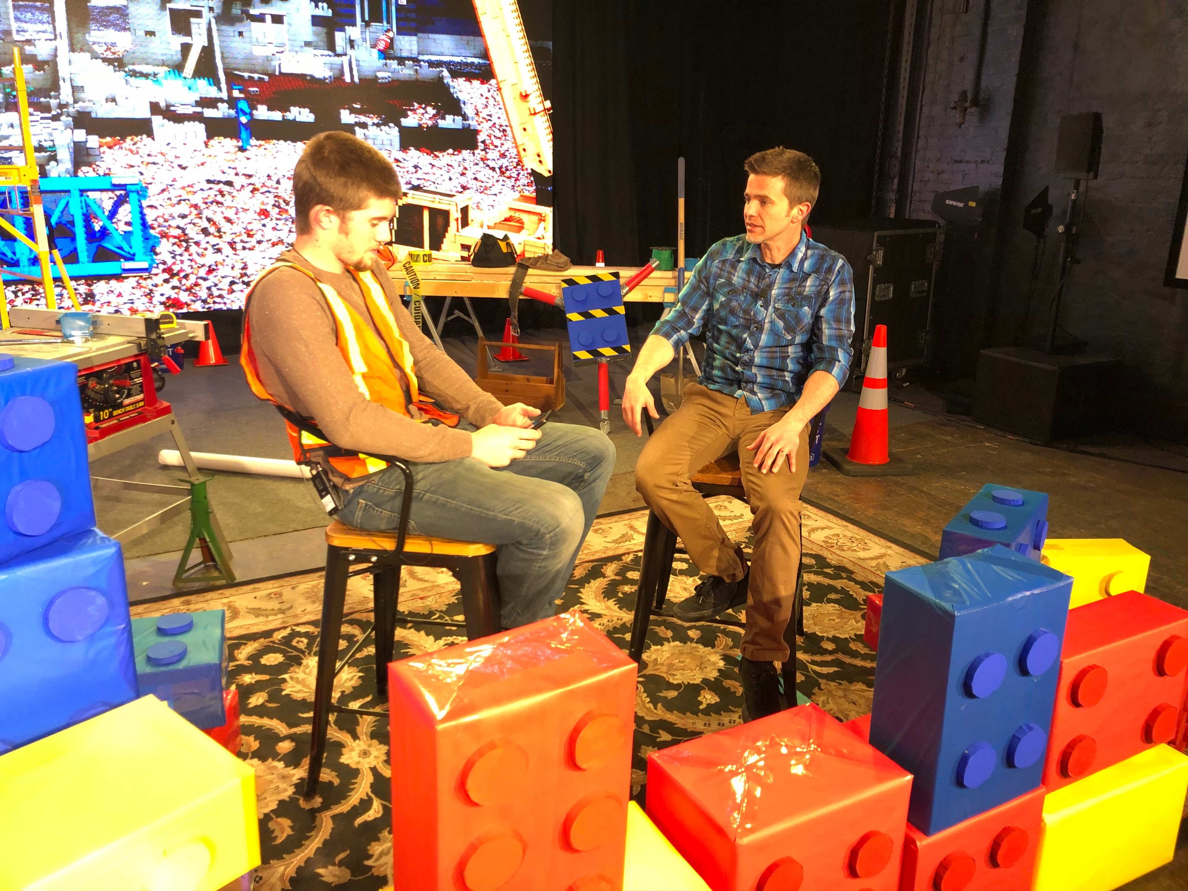 Two men sit on chairs surrounded by large, colorful blocks in a room set up for a video shoot.