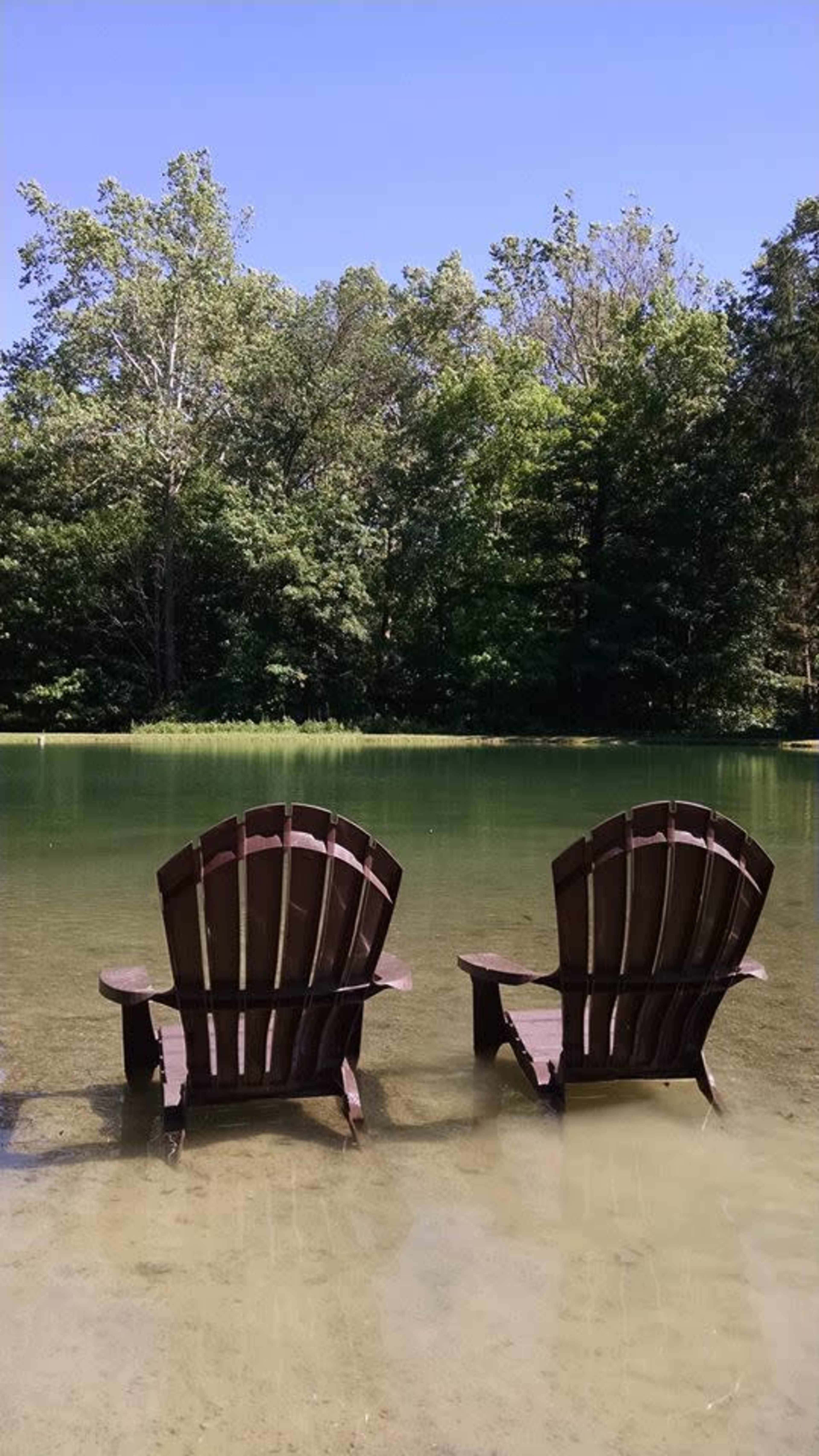 Two wooden Adirondack chairs sit partially submerged in calm water with a forested area in the background.