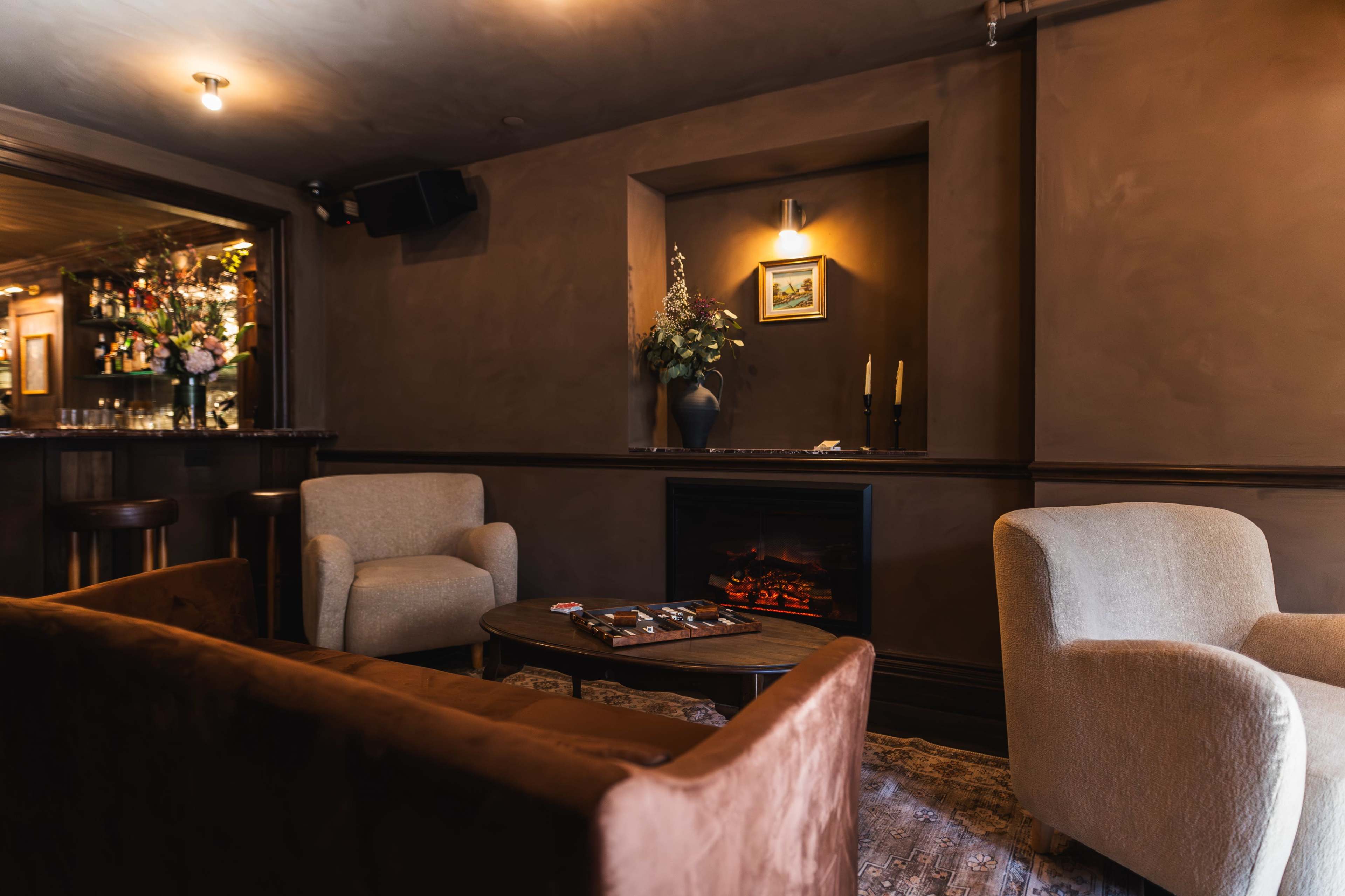 A cozy seating area with two armchairs, a small table, and a faux fireplace, set in a dimly lit room decorated in warm tones.